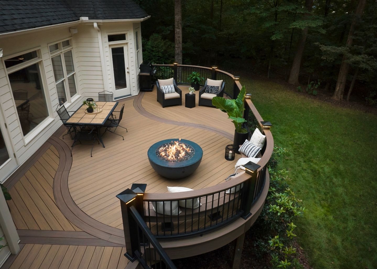Terrasse en bois incurvée avec sièges extérieurs et foyer à côté d'une maison, donnant sur une cour arrière gazonnée.