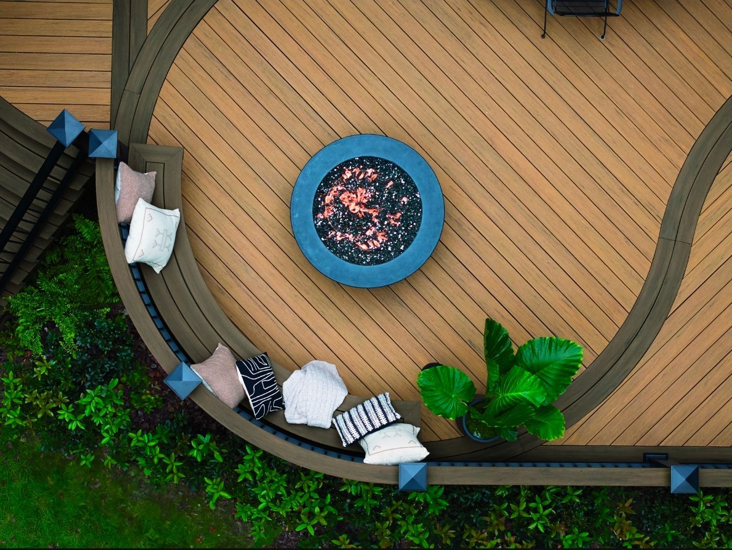 Vue aérienne d'une terrasse en bois avec un foyer rond, des bancs incurvés et des plantes vertes.