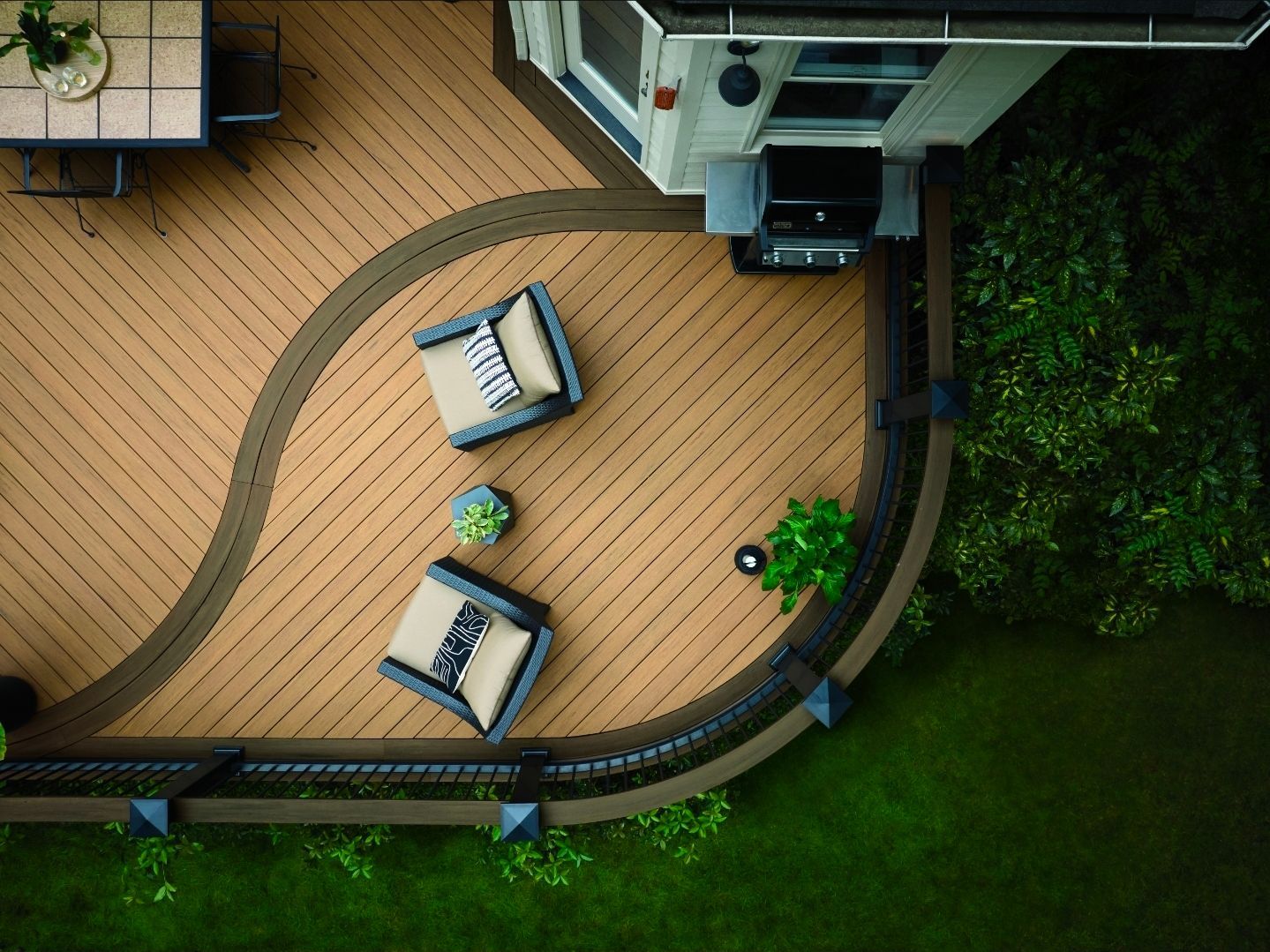 Vue aérienne d'une terrasse en bois incurvée avec deux chaises, une petite table et un barbecue, à côté d'une pelouse verte et d'arbustes.