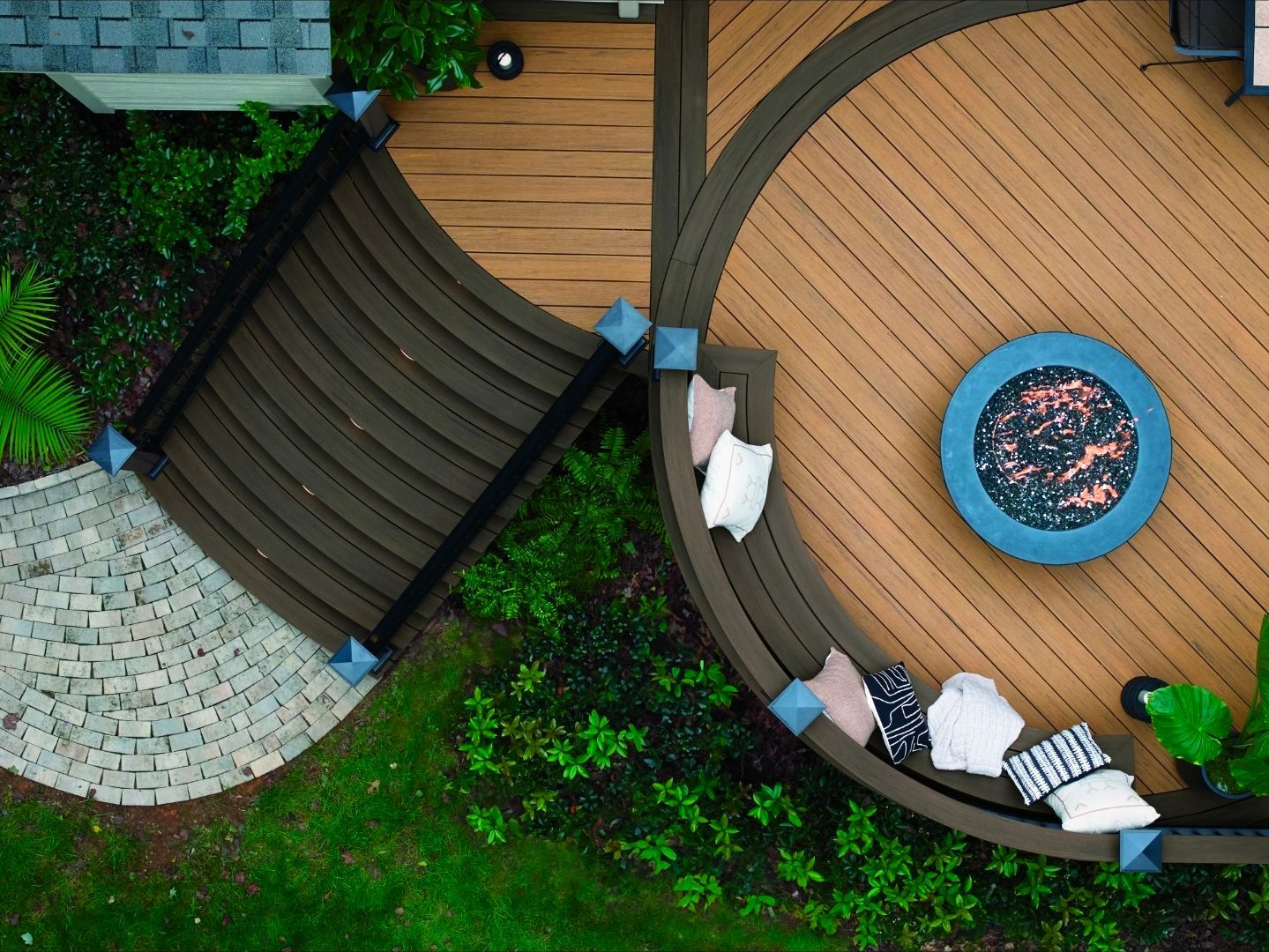 Vue aérienne d'une terrasse en bois incurvée avec un foyer rond, des escaliers, des plantes en pot et une terrasse en pierre