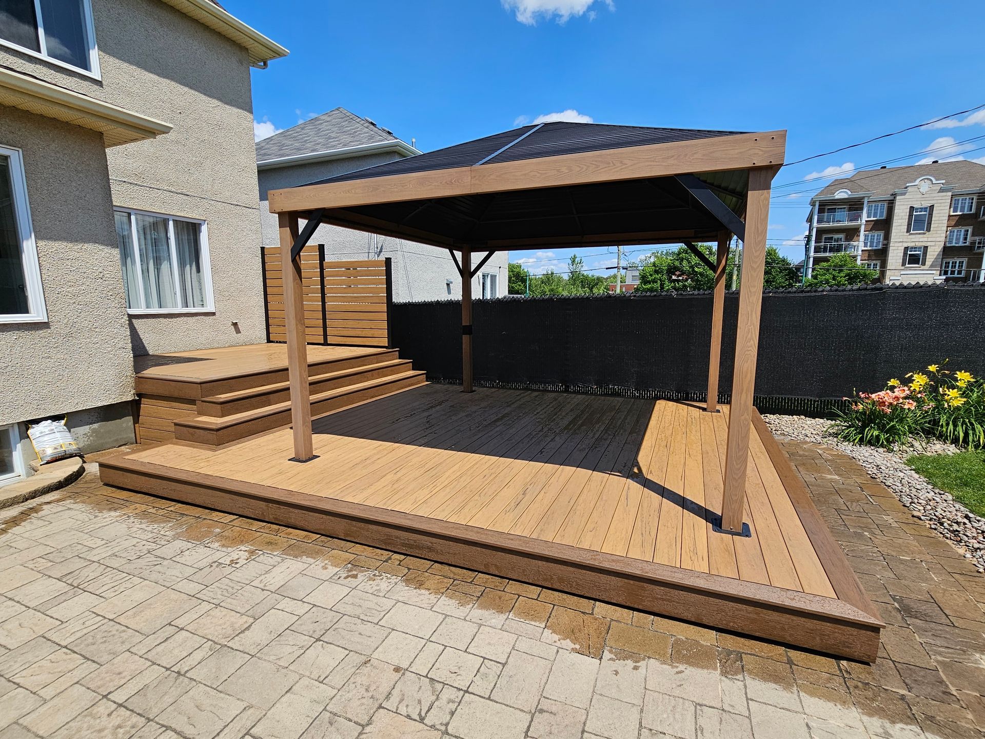 Terrasse arrière avec pergola en bois et marches jouxtant une maison en stuc, bordée de pavés et de fleurs