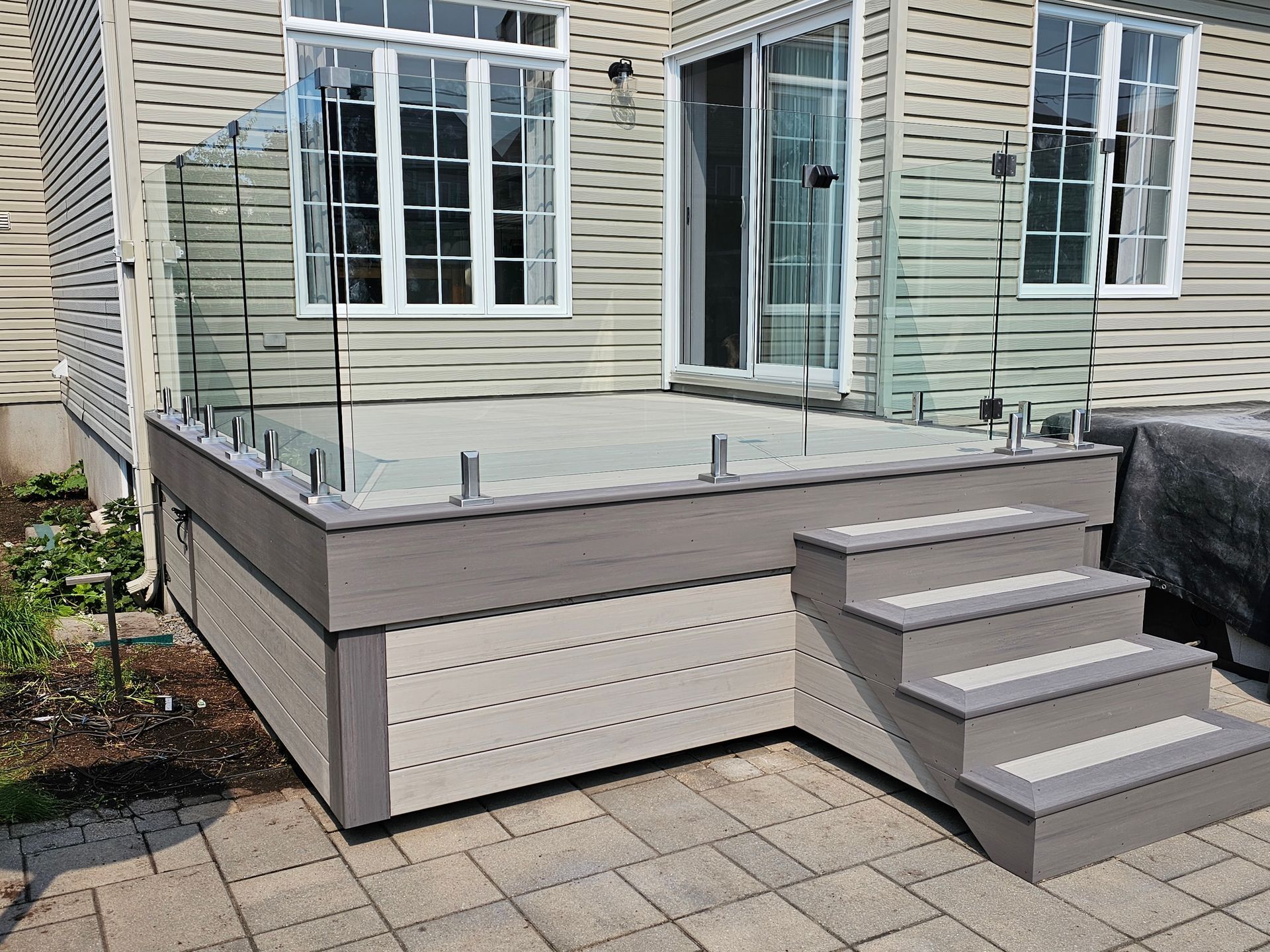 Terrasse arrière avec marches grises et balustrade en verre, attenante à la maison, patio pavé en contrebas.