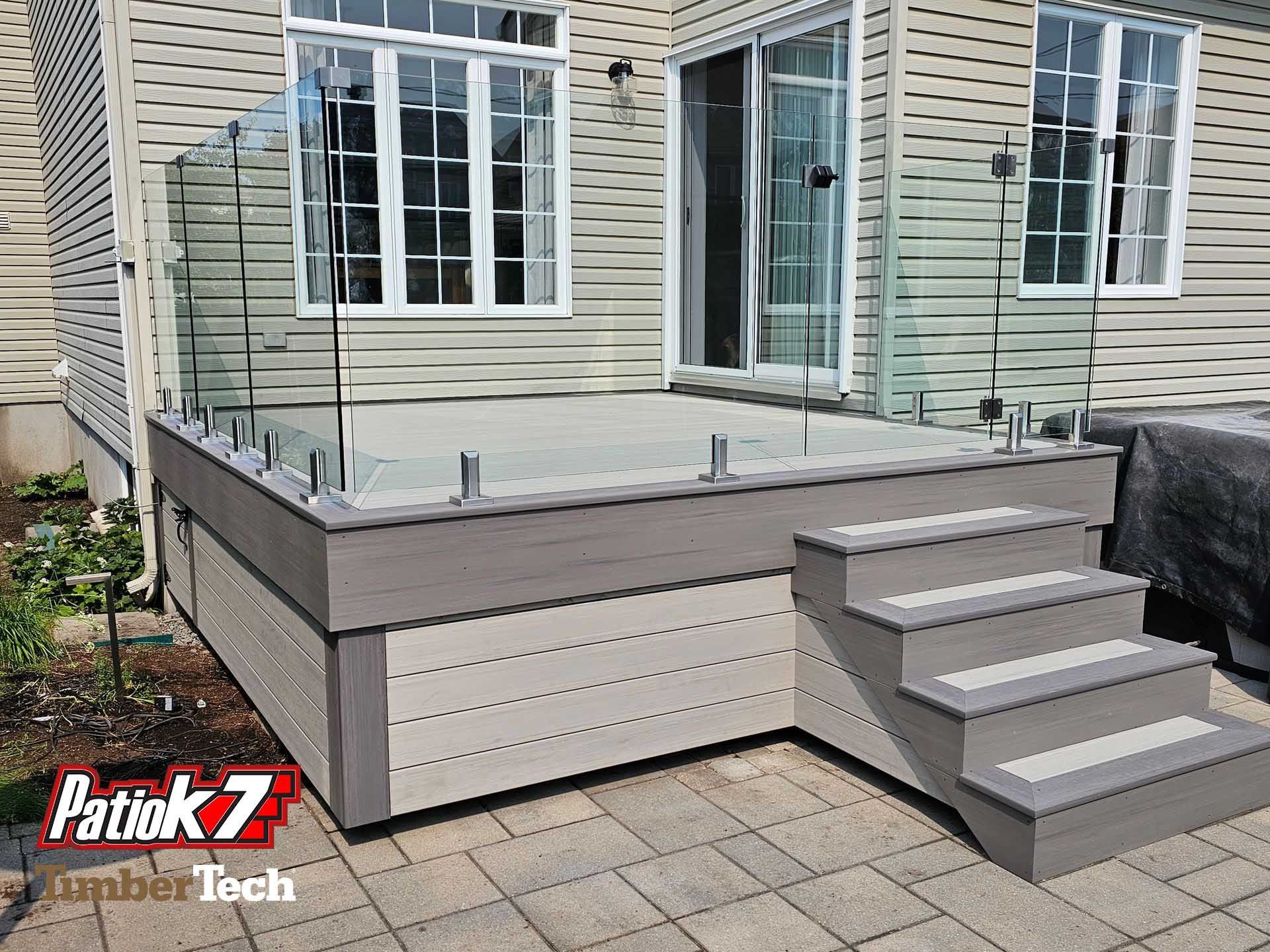 Terrasse arrière avec marches grises et balustrade en verre, attenante à la maison, patio pavé en contrebas.