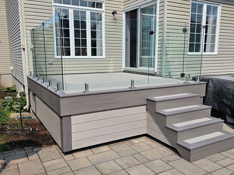 Terrasse arrière avec marches grises et balustrade en verre, attenante à la maison, patio pavé en contrebas.