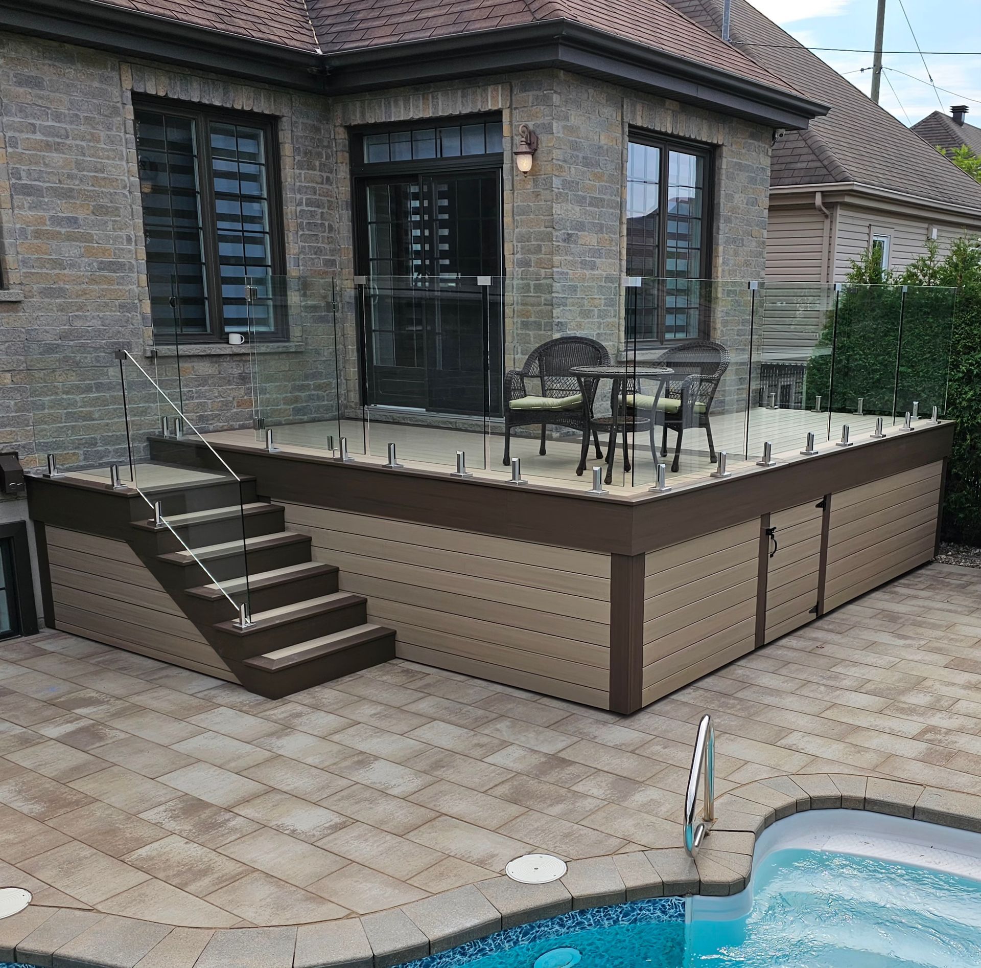 Terrasse surélevée attenante à une maison en briques, avec escalier, table et chaises, à côté d'une piscine.