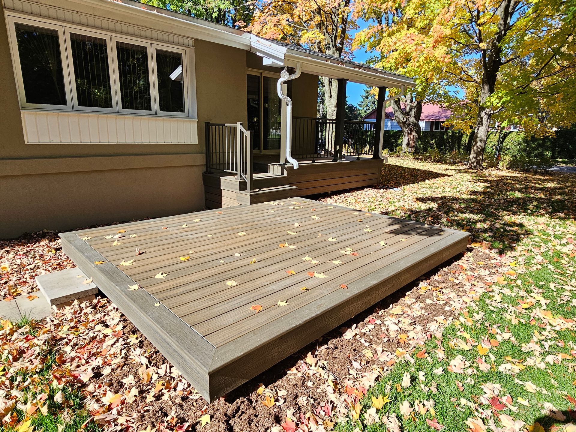 Terrasse en bois jouxtant une maison beige, entourée de feuilles mortes d'automne et d'arbres dans un jardin.