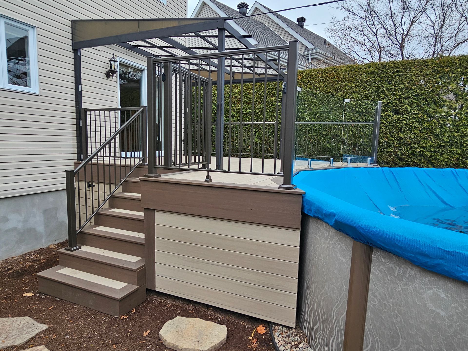 Escalier de terrasse arrière avec structure de pergola, bardage beige et piscine recouverte de bleu à côté d'une haie