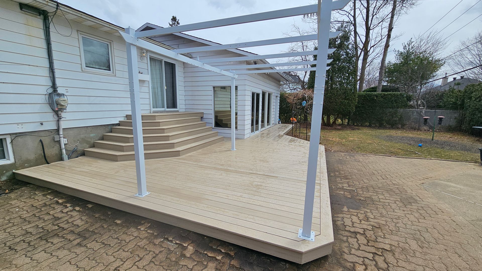 Terrasse en construction dans le jardin, avec poteaux et escaliers blancs, à côté d'une maison.
