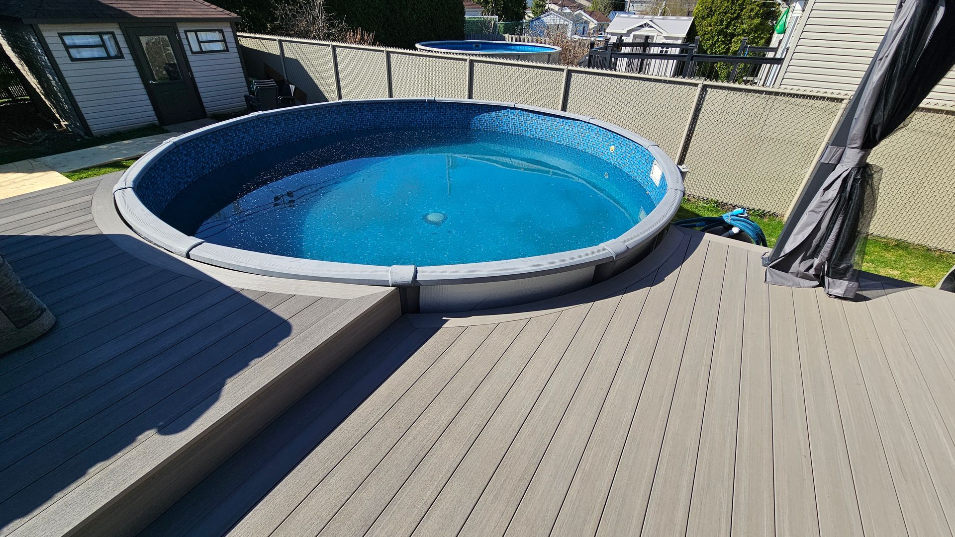 Piscine hors sol bleue sur une terrasse en bois dans une cour arrière clôturée, avec un abri de jardin et un coin salon couvert à proximité.