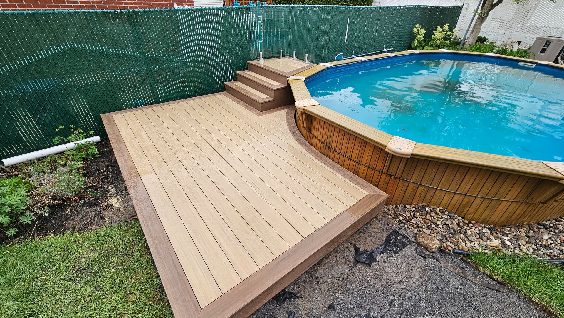 Terrasse en bois jouxtant une piscine hors sol avec des marches et une clôture verte assurant l'intimité dans une cour arrière.