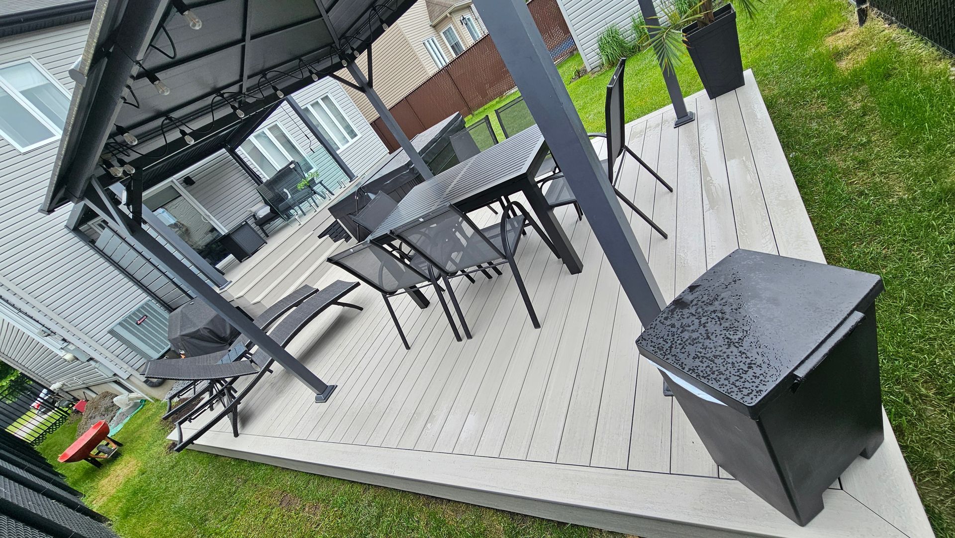 Terrasse extérieure couverte avec table et chaises noires sur une terrasse en bois bordant la pelouse.