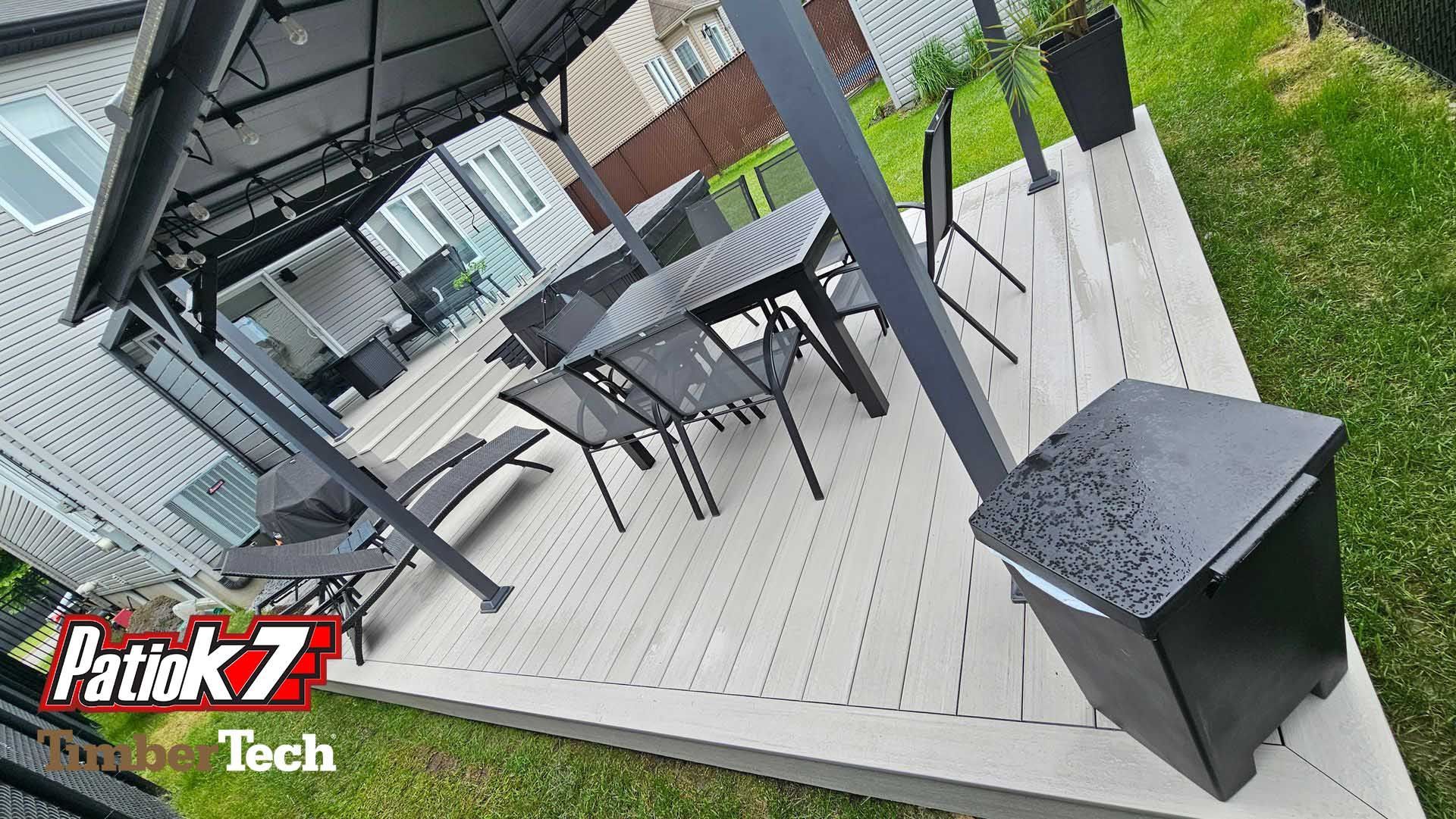 Terrasse extérieure couverte avec table et chaises noires sur une terrasse en bois bordant la pelouse.