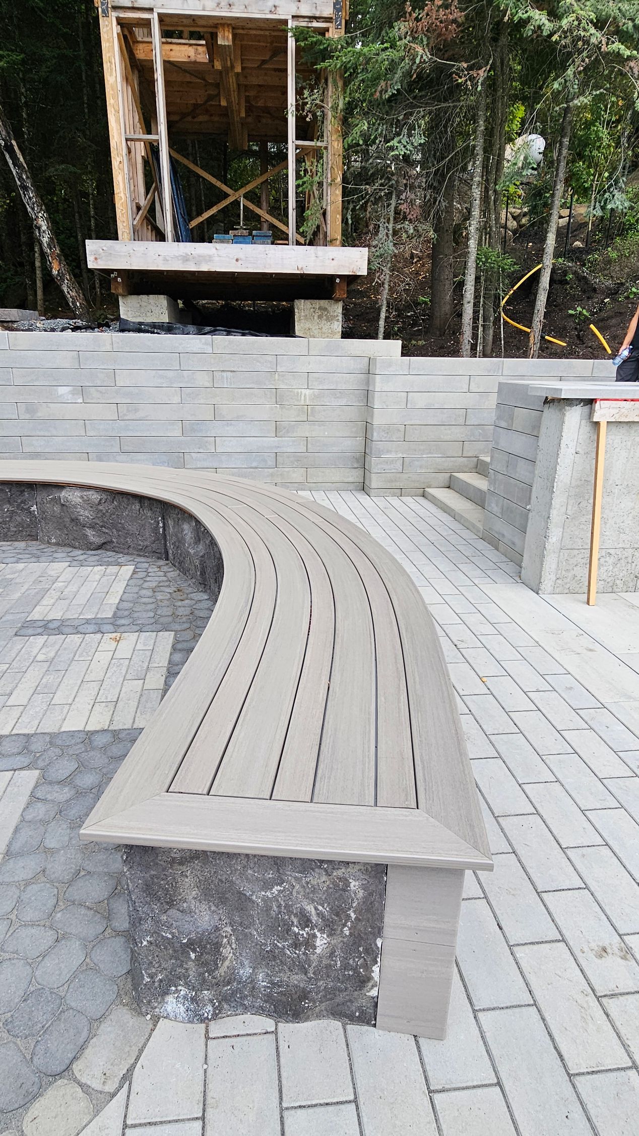 Banc en bois courbé dans un jardin extérieur pavé, avec des arbres et une petite structure en arrière-plan.