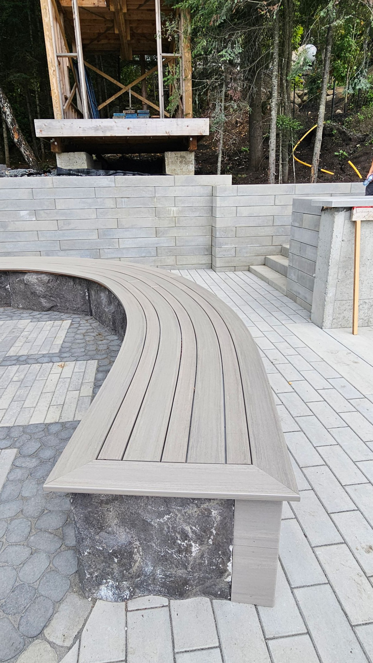 Banc en bois courbé sur une terrasse pavée, à côté d'un muret en pierre, avec des arbres et une petite construction en arrière-plan.