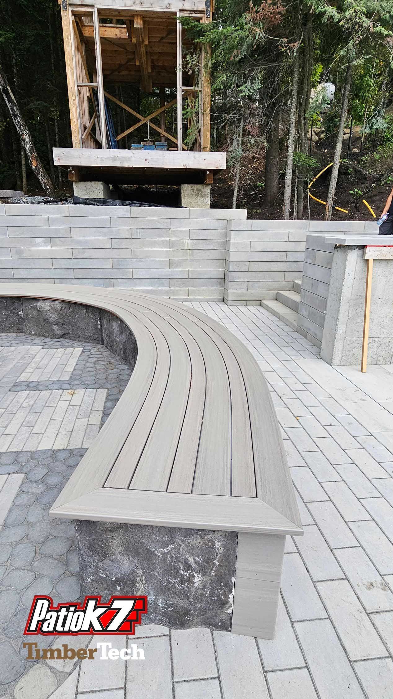Banc en bois courbé sur une terrasse pavée, à côté d'un muret en pierre, avec des arbres et une petite construction en arrière-plan.