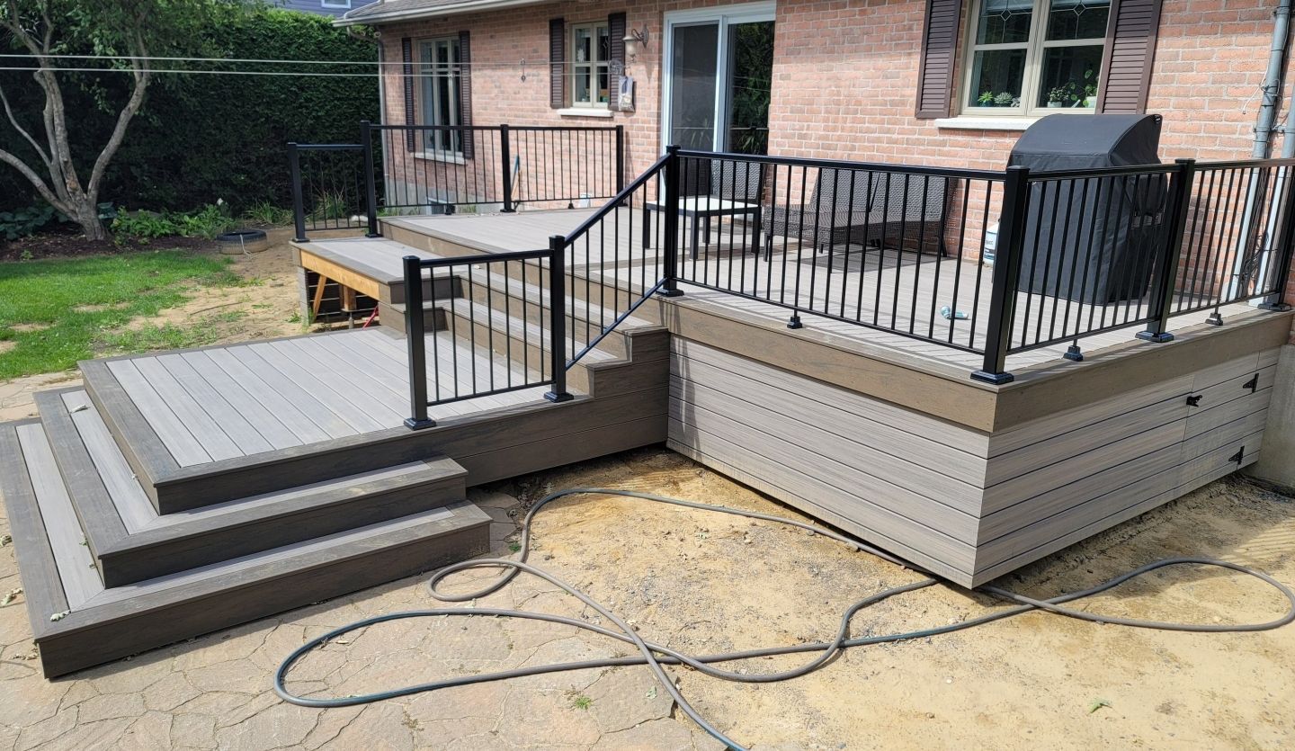 Terrasse arrière avec rambardes noires, marches grises et plinthes beiges, jouxtant une maison en briques.