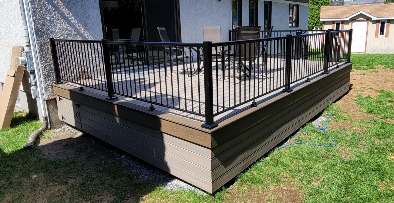 Terrasse surélevée dans la cour arrière, avec rambarde noire et plinthe beige, jouxtant une maison et une pelouse.