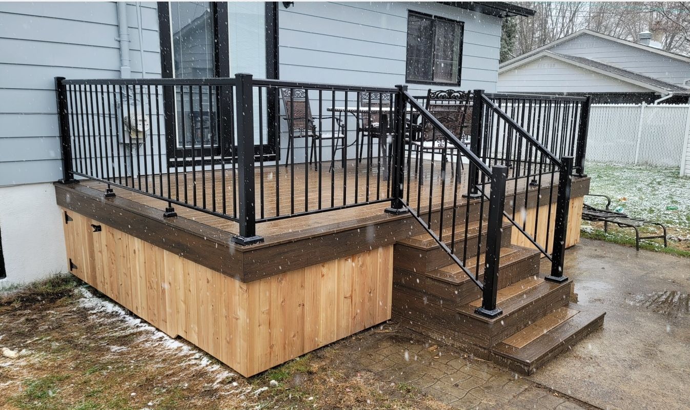Terrasse en bois avec rambarde et marches en métal noir, jouxtant une maison sur une terrasse en béton humide.