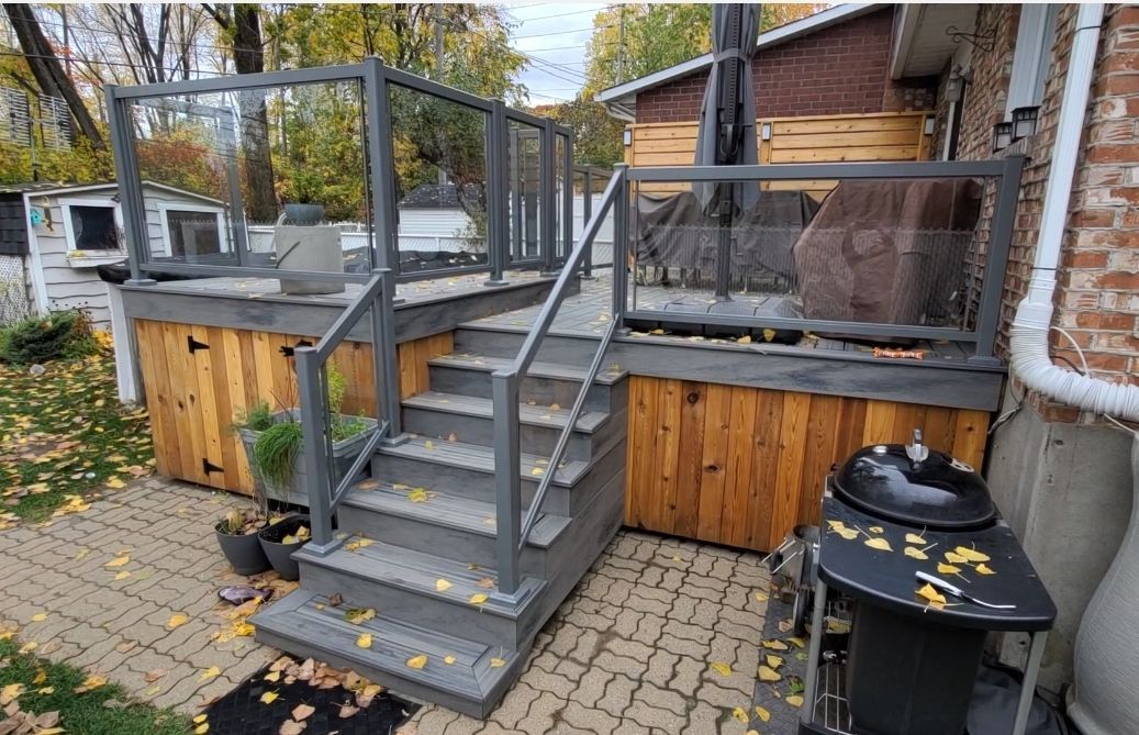 Terrasse en bois avec escalier et rambarde en métal, maison en briques et barbecue sur un patio pavé jonché de feuilles mortes.