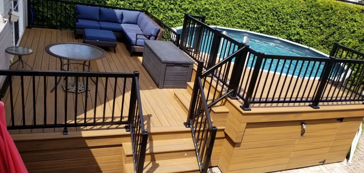 Terrasse avec rambarde noire, canapé d'angle bleu d'extérieur, table ronde et jacuzzi derrière une haie.