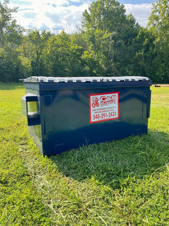 Dark blue dumpster on green grass with trees in the background. Has a sticker with a phone number.