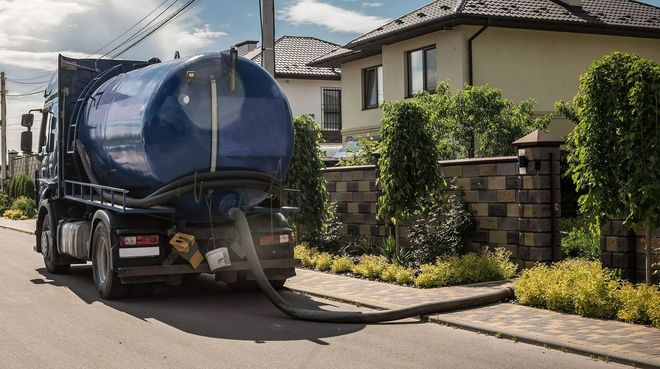 Blue septic tank truck parked in front of a house, hose extended to the curb.
