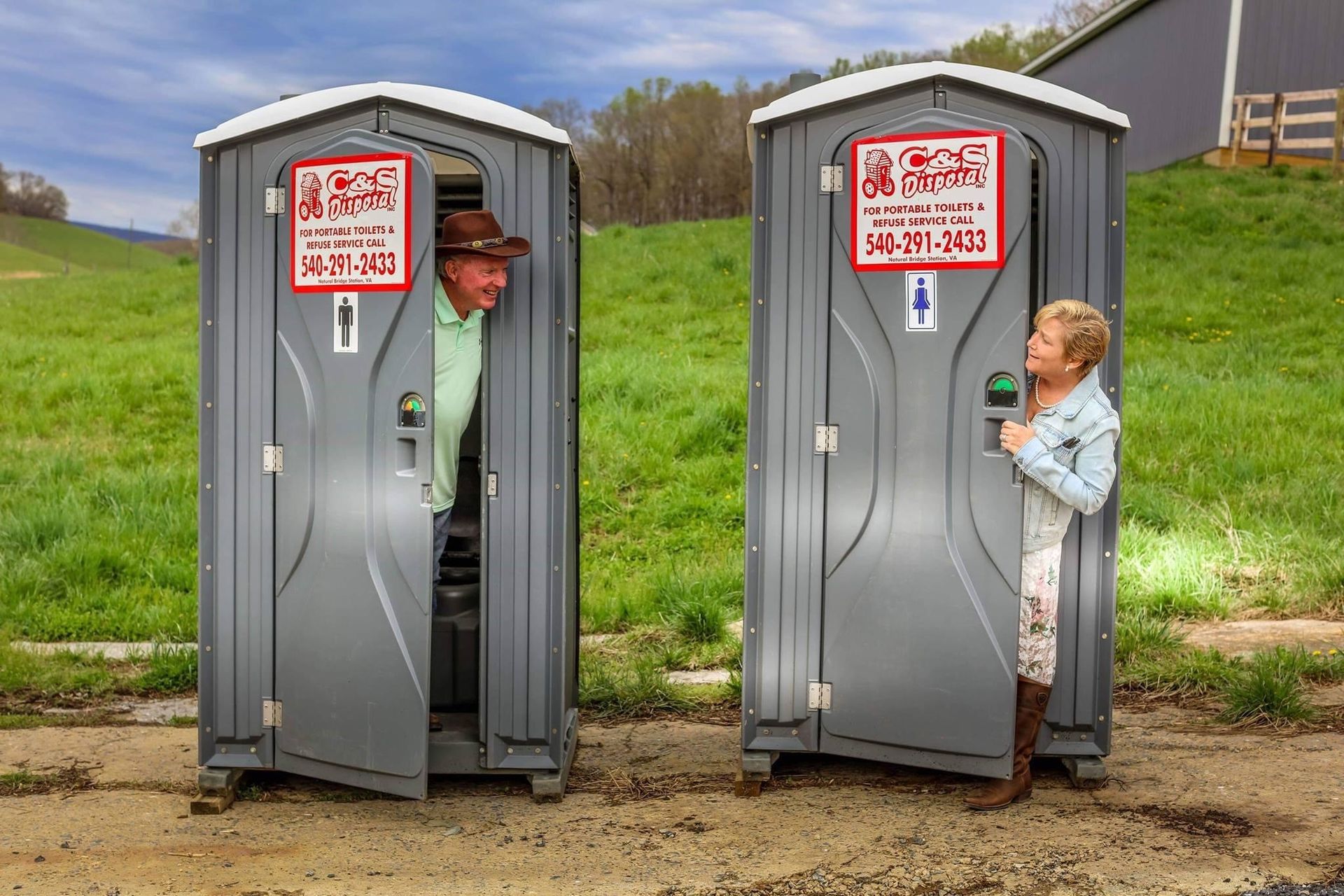 A man in a cowboy hat and a woman smile from portable toilets in a rural setting.