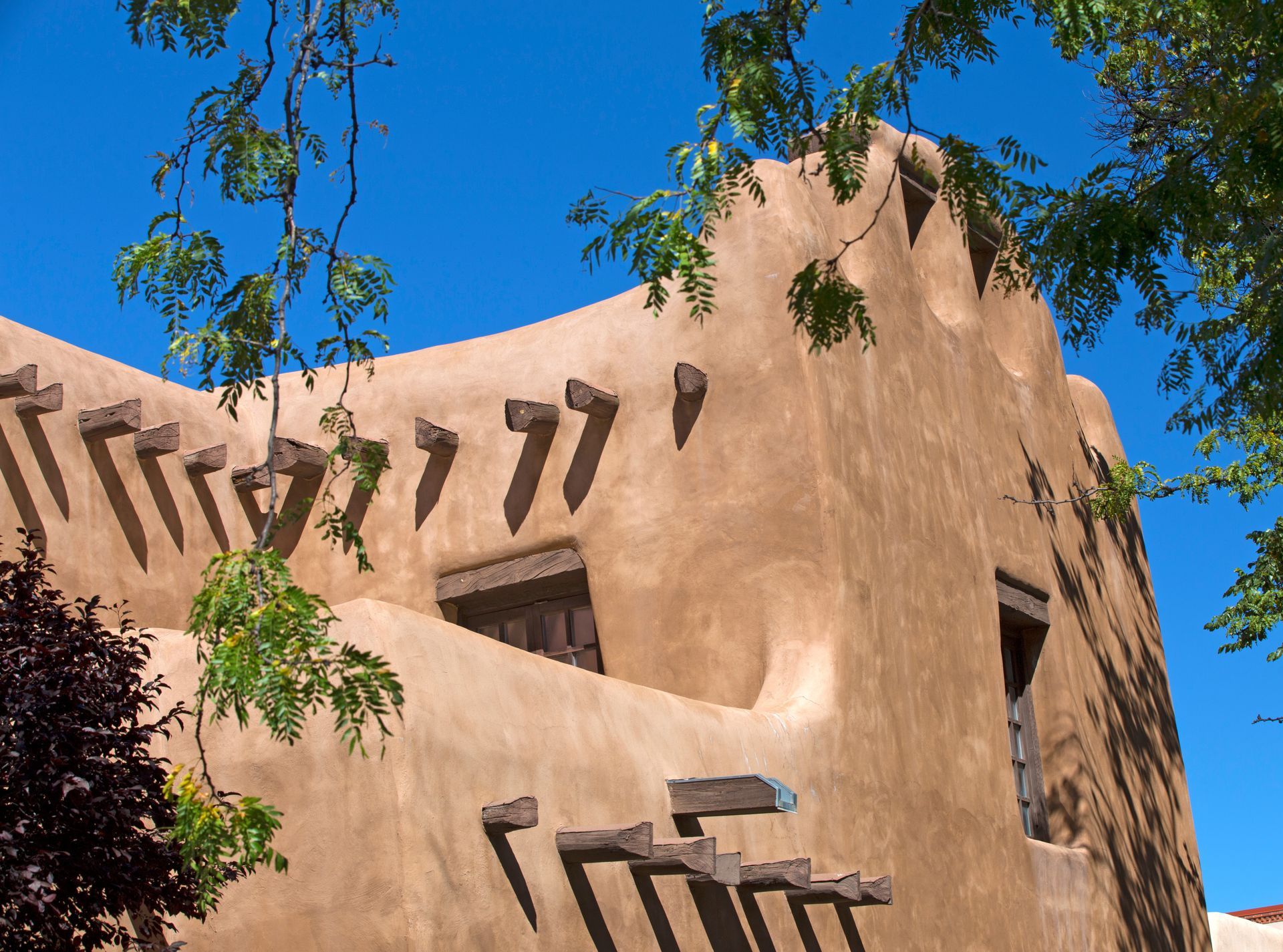 Adobe-style stucco building with angular windows and tree branches against a bright blue sky Adobe-style stucco building with angular windows and tree branches against a bright blue sky