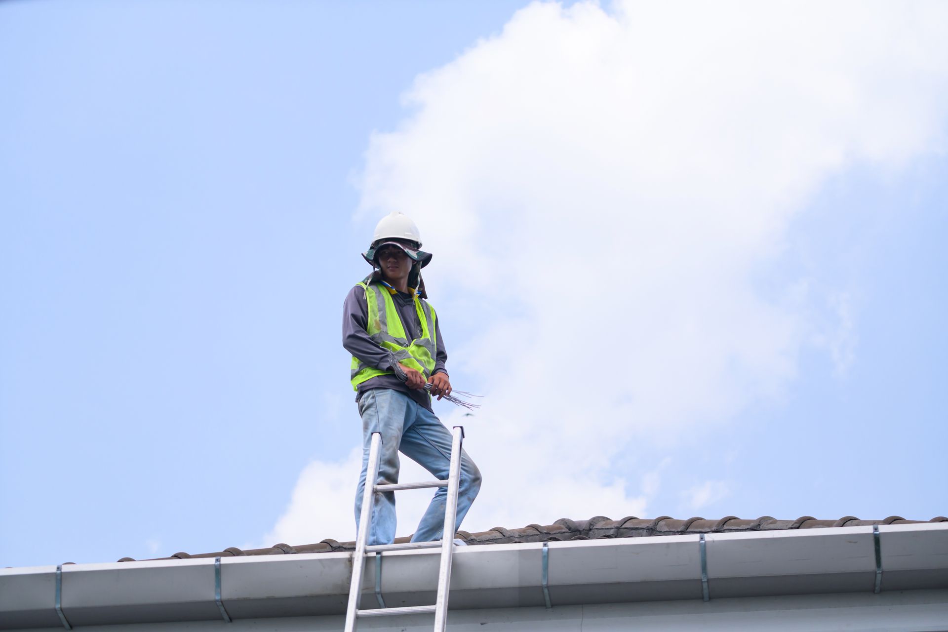 A roofer in a protective hat and safety vest on a tile roof beside a ladder.