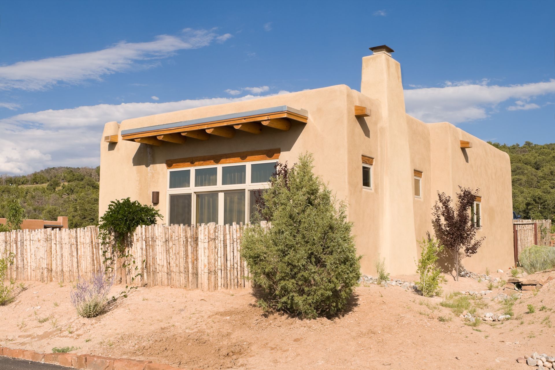 Desert adobe house with tan stucco walls, wooden fence, and shrubs under a blue sky Desert adobe house with tan stucco walls, wooden fence, and shrubs under a blue sky