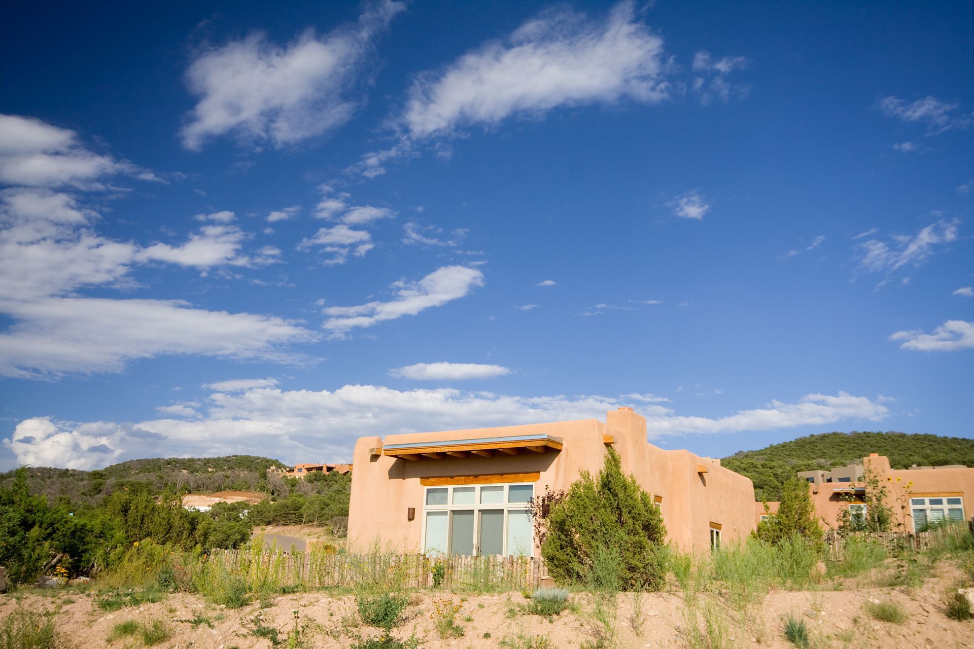 Desert adobe house with tan stucco walls, wooden fence, and shrubs under a blue sky Small peach-colored house in a grassy field under a bright blue sky with clouds