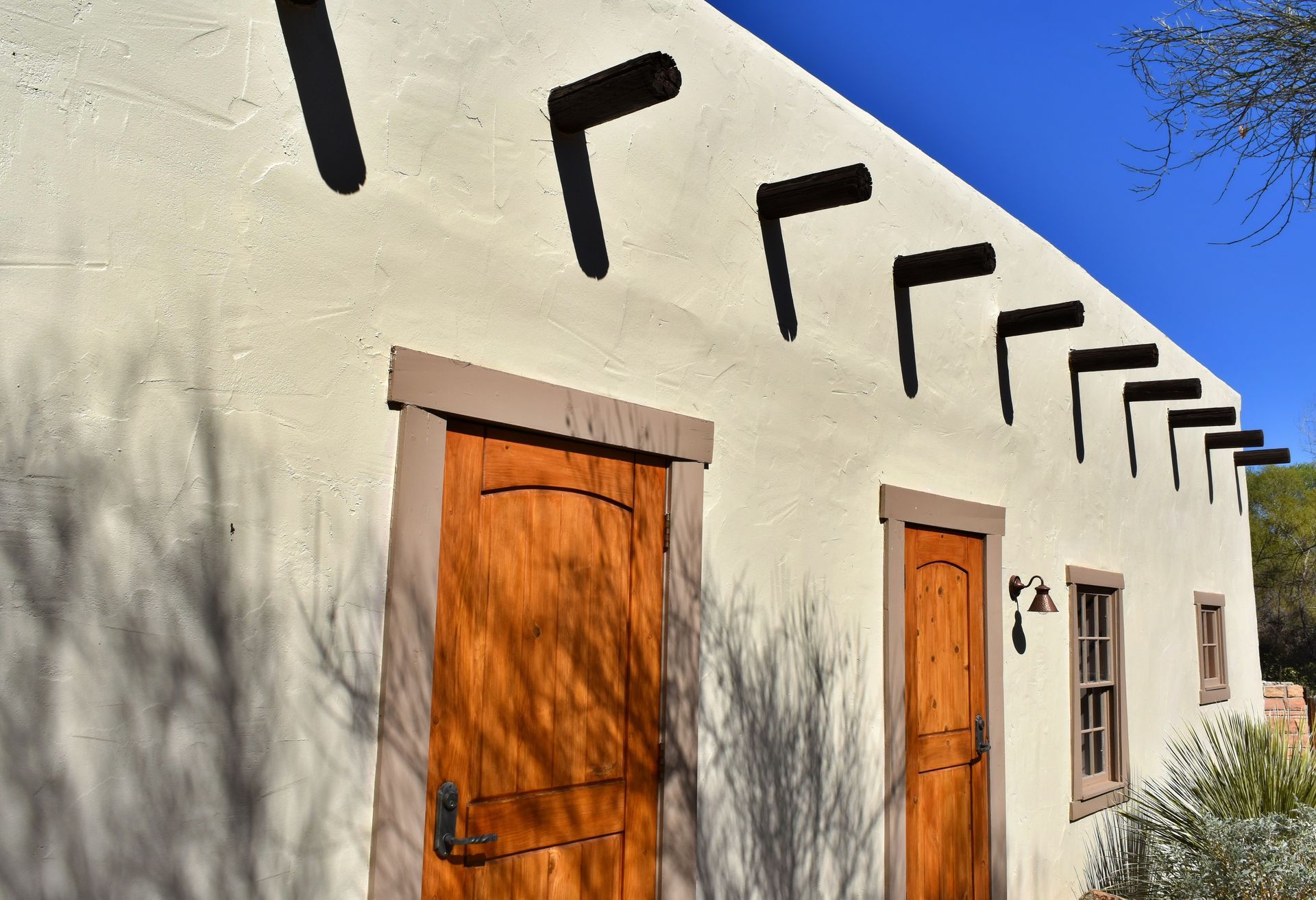 White stucco building with wooden doors and black roof beams casting shadows in sunlight