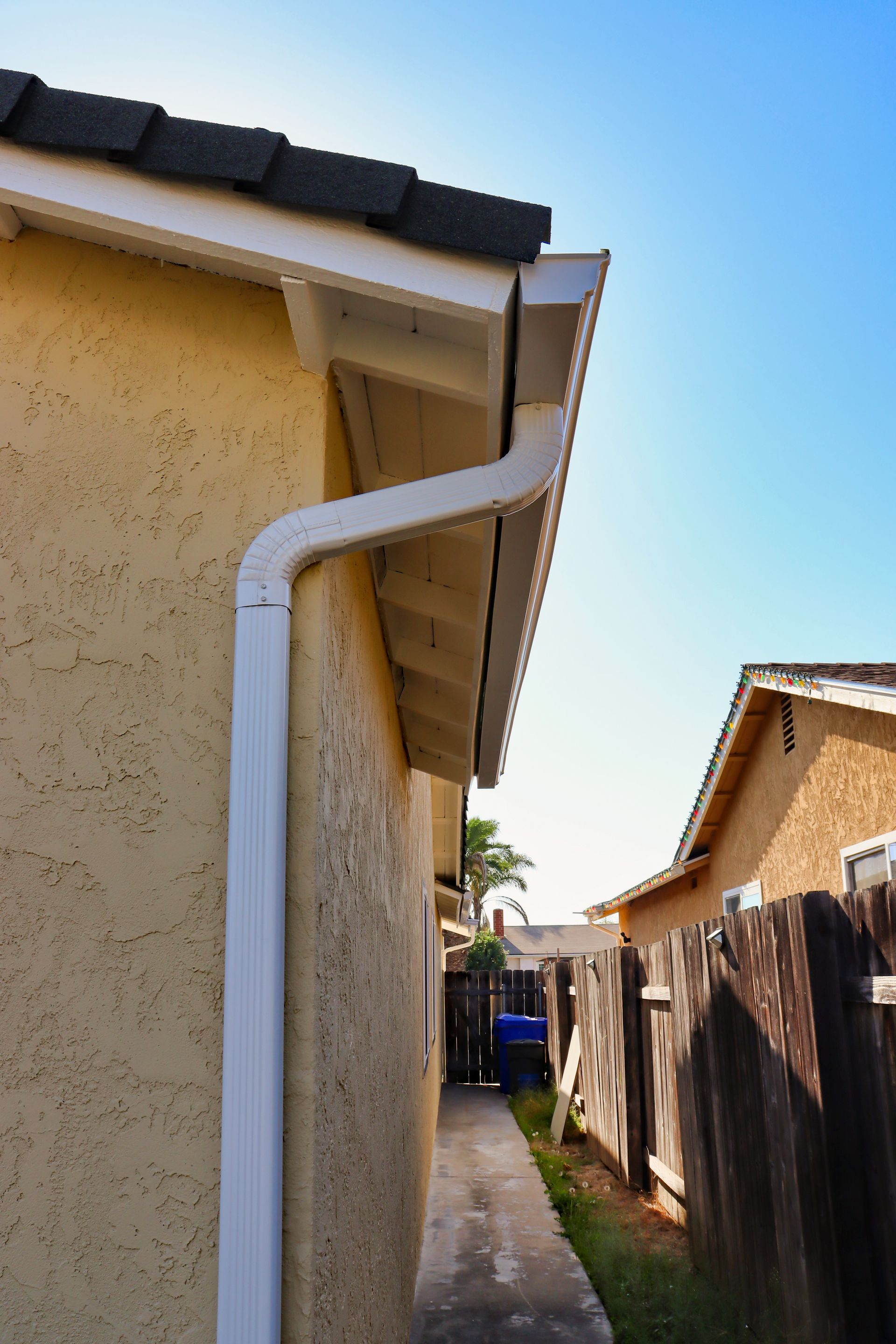 Narrow side yard between tan houses, with a white downspout and blue sky overhead.