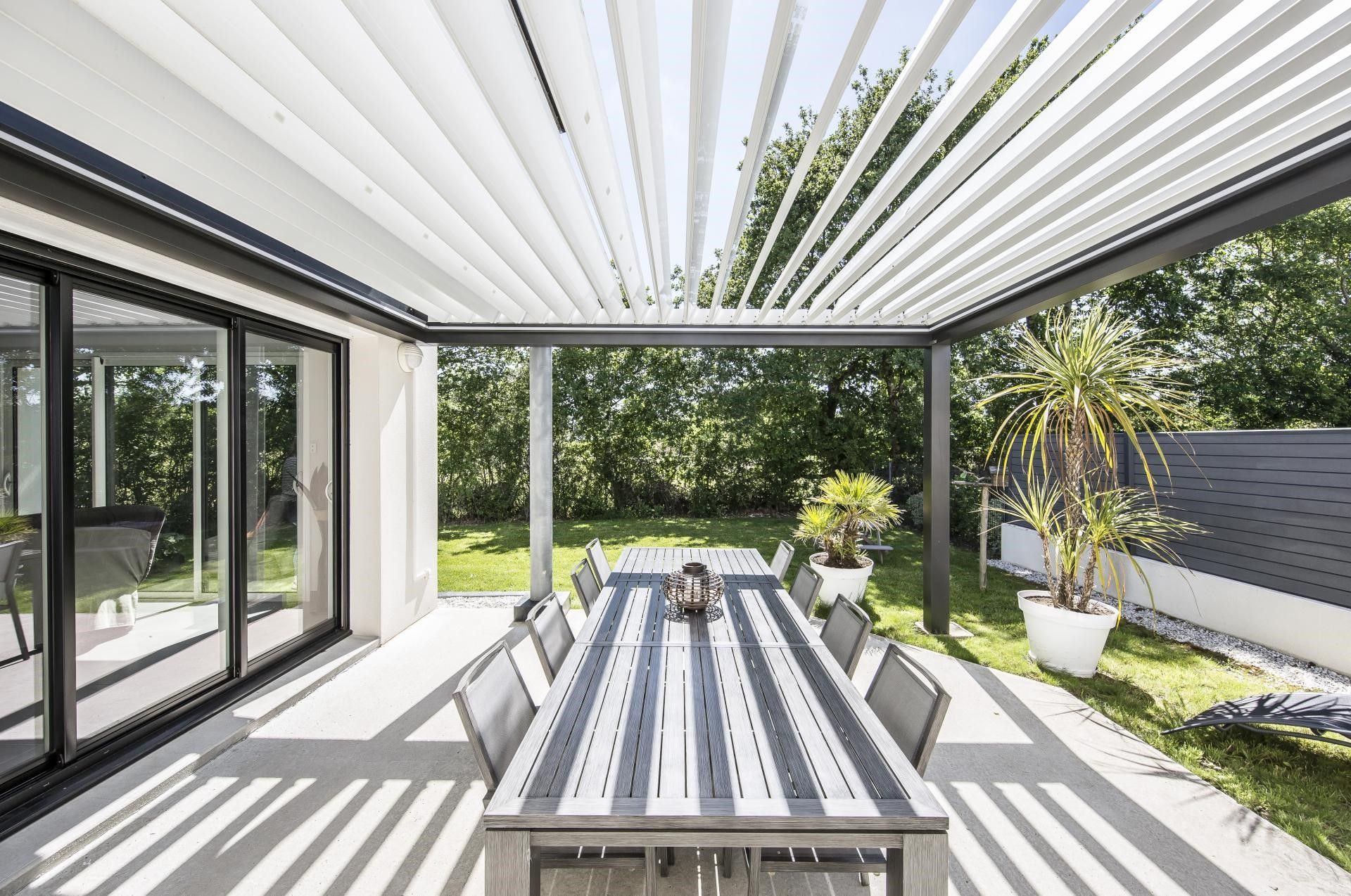 Patio coperto con tavolo da pranzo all'aperto, lamelle bianche per pergola, porte scorrevoli in vetro e vista sul giardino.