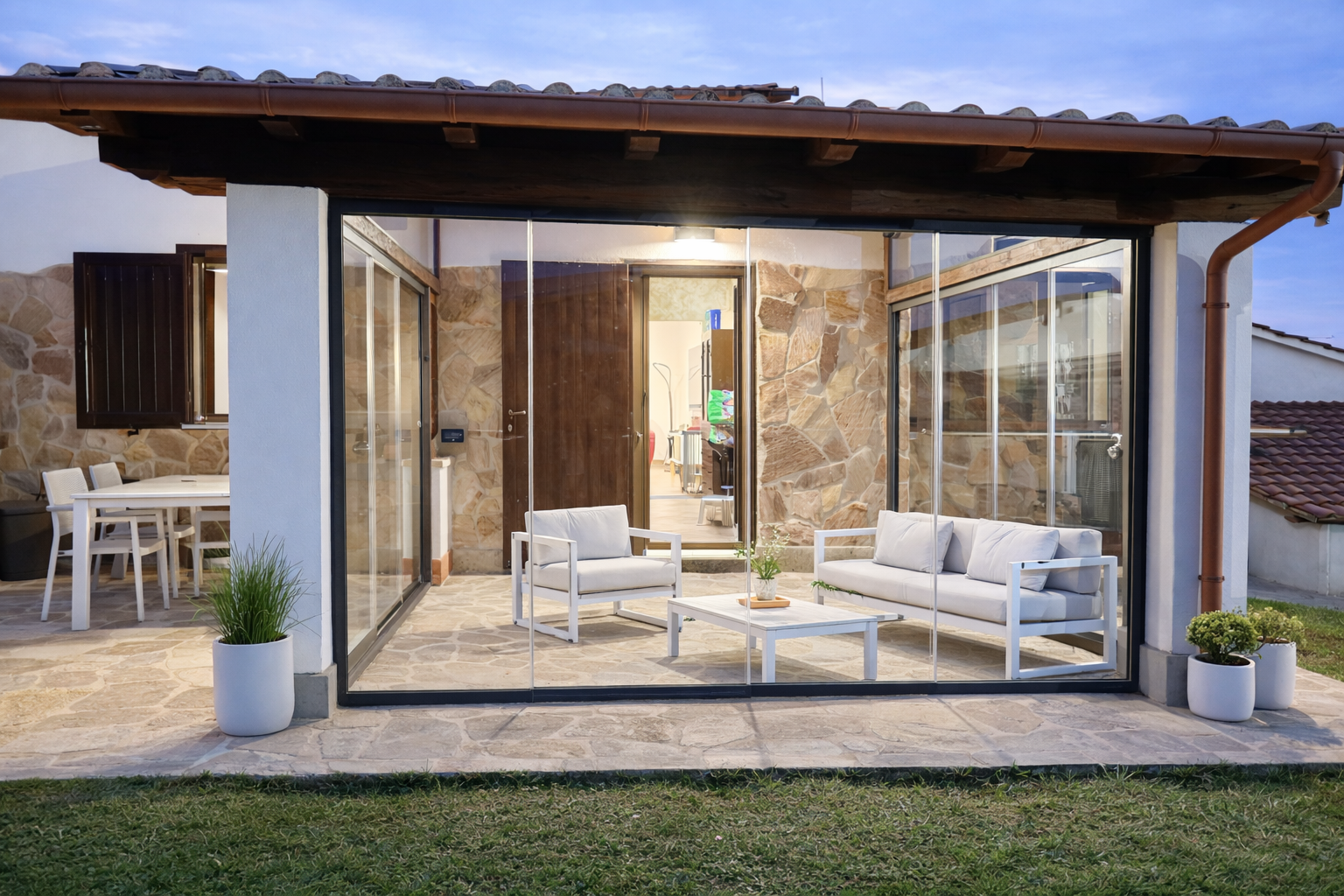 Patio moderno con pareti in vetro, mobili da esterno bianchi e luci interne calde al crepuscolo.