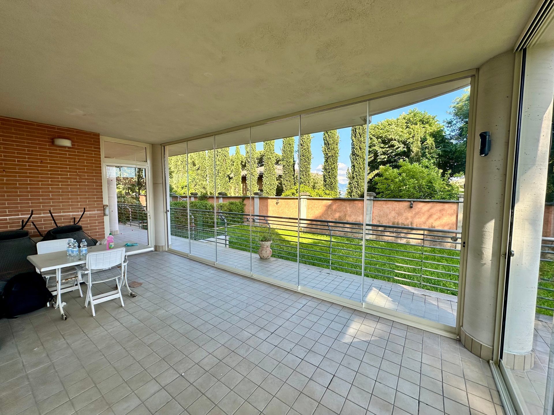 Patio coperto con tavolo e sedie bianchi, pavimento piastrellato e muro in mattoni che si affaccia su un prato e sugli alberi.