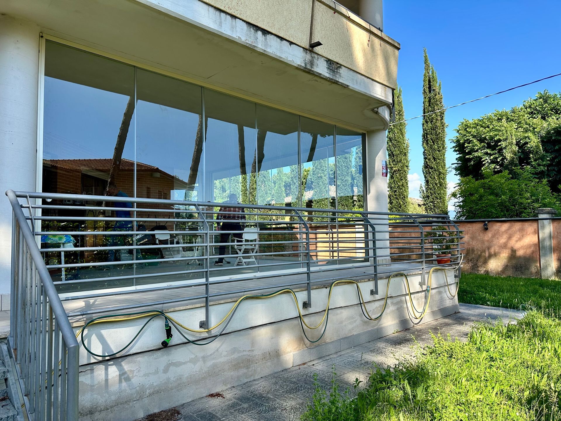 Patio di una casa moderna con finestre in vetro, ringhiera in metallo e un piccolo giardino lungo il vialetto.