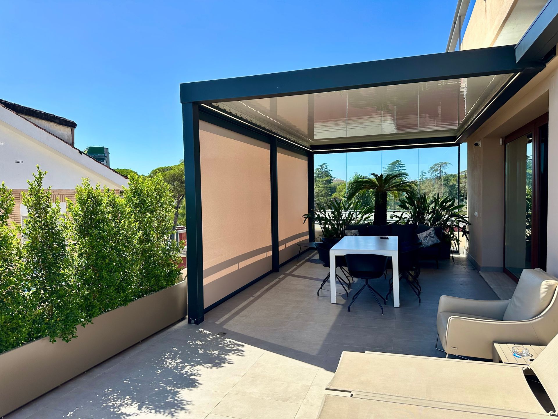 Patio coperto con tenda da sole con struttura nera, tavolo bianco e sedia a sdraio con vista su un giardino soleggiato.