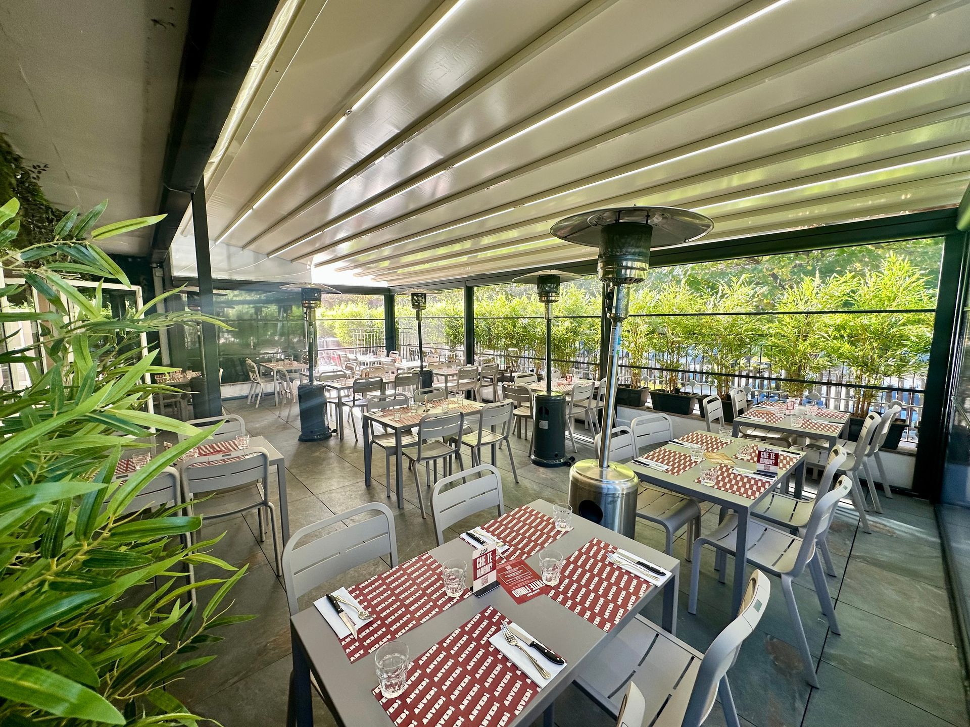 Terrazza ristorante all'aperto con tavoli, sedie e tenda a righe, circondata dal verde.