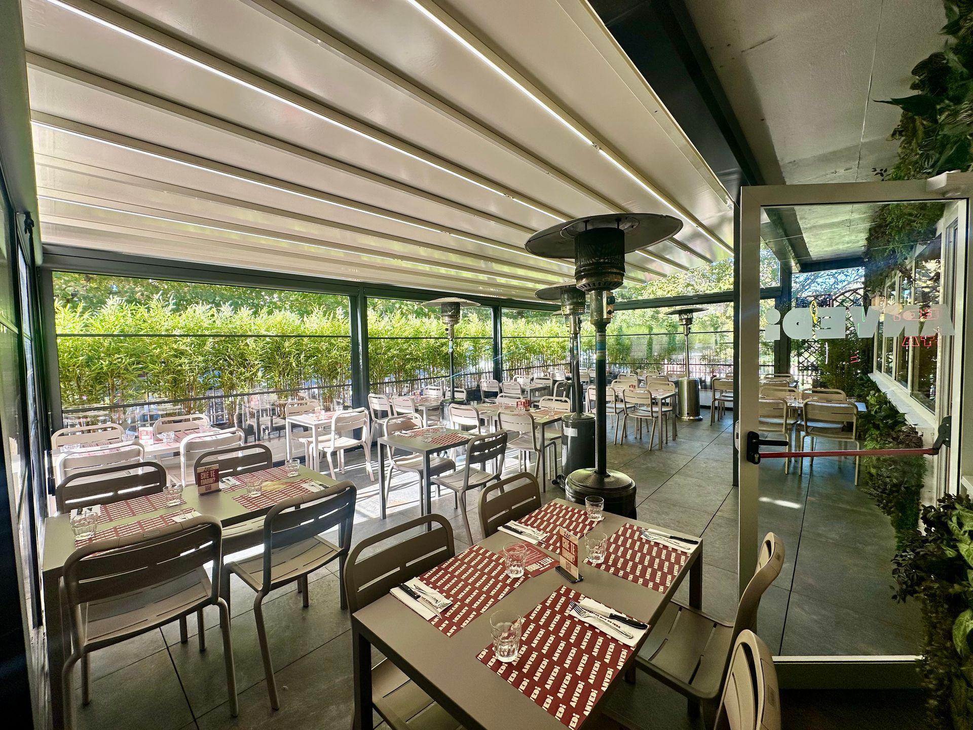 Terrazza esterna del caffè con tende a righe, tavoli e sedie vuoti e alberi oltre la ringhiera di vetro.