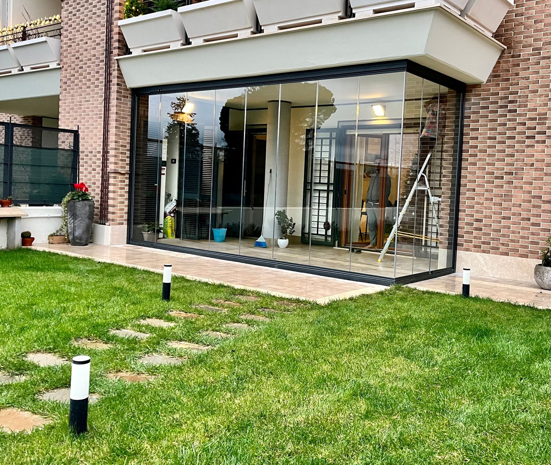 Patio vetrato con finestre incorniciate di nero, adiacente a un edificio in mattoni e con prato verde antistante.