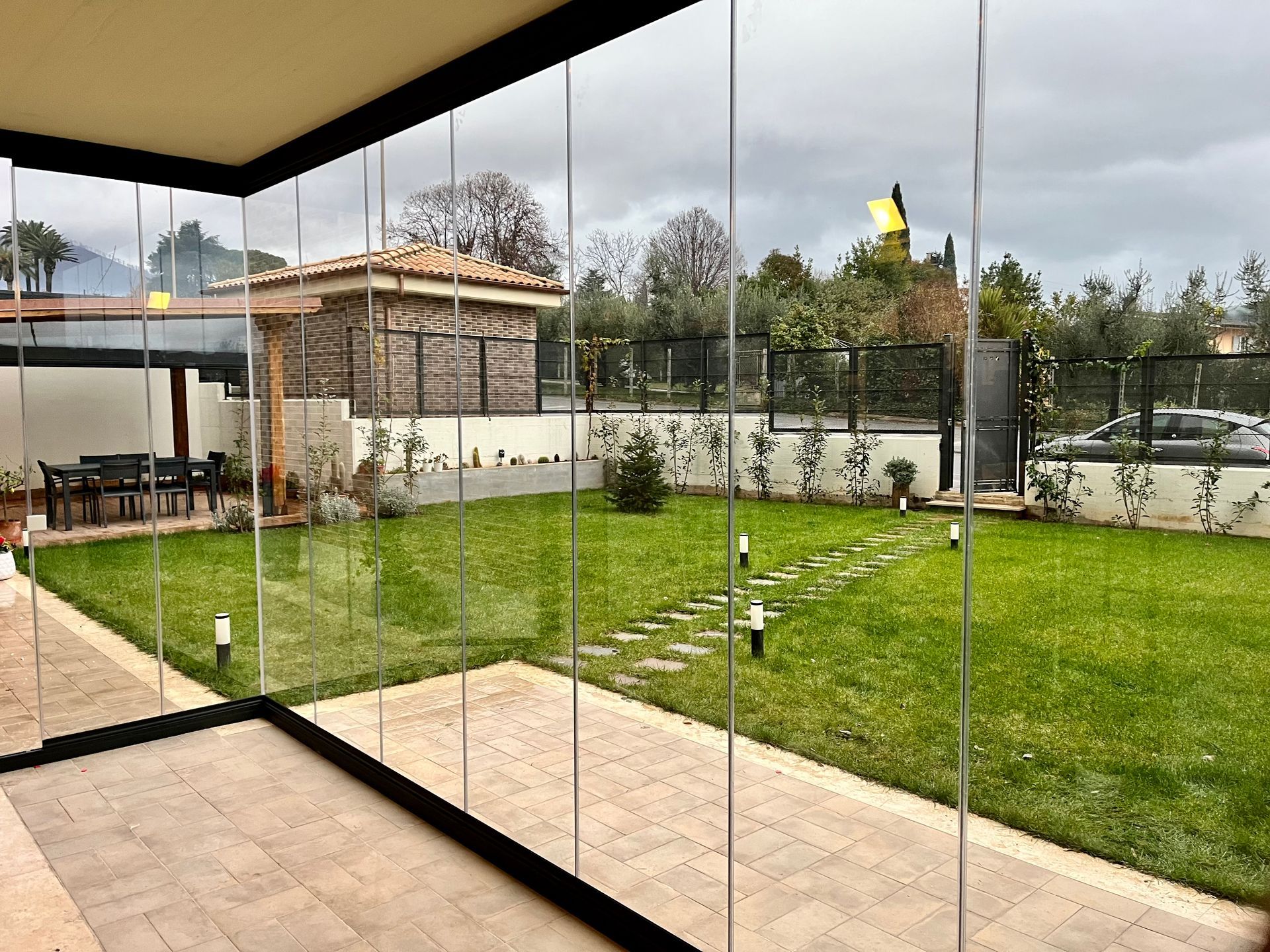 Patio con pareti in vetro affacciato su un prato verde e un giardino sotto un cielo nuvoloso.