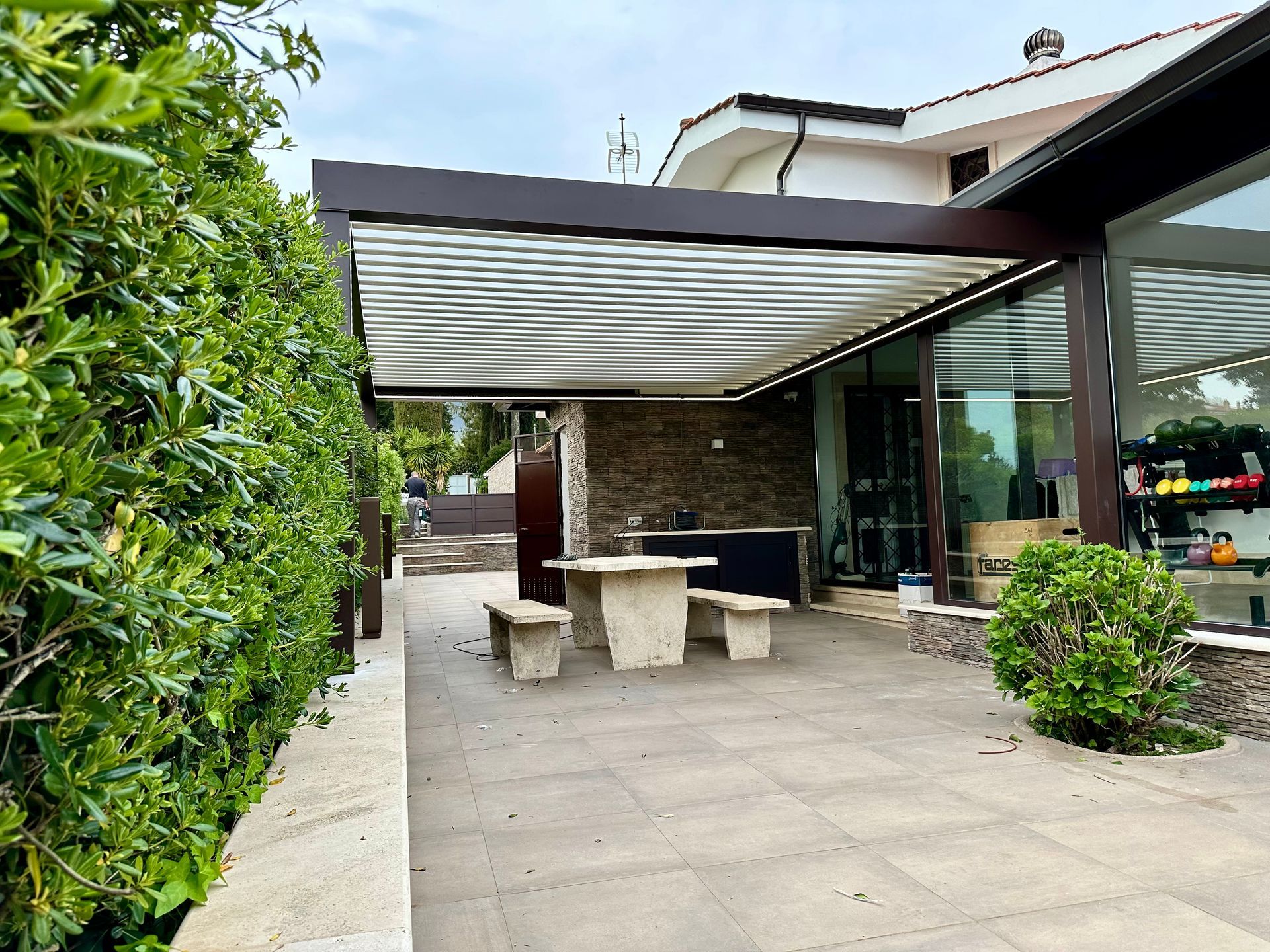 Patio con tavolo e panchine in pietra sotto un pergolato, accanto a una casa moderna e al giardino.