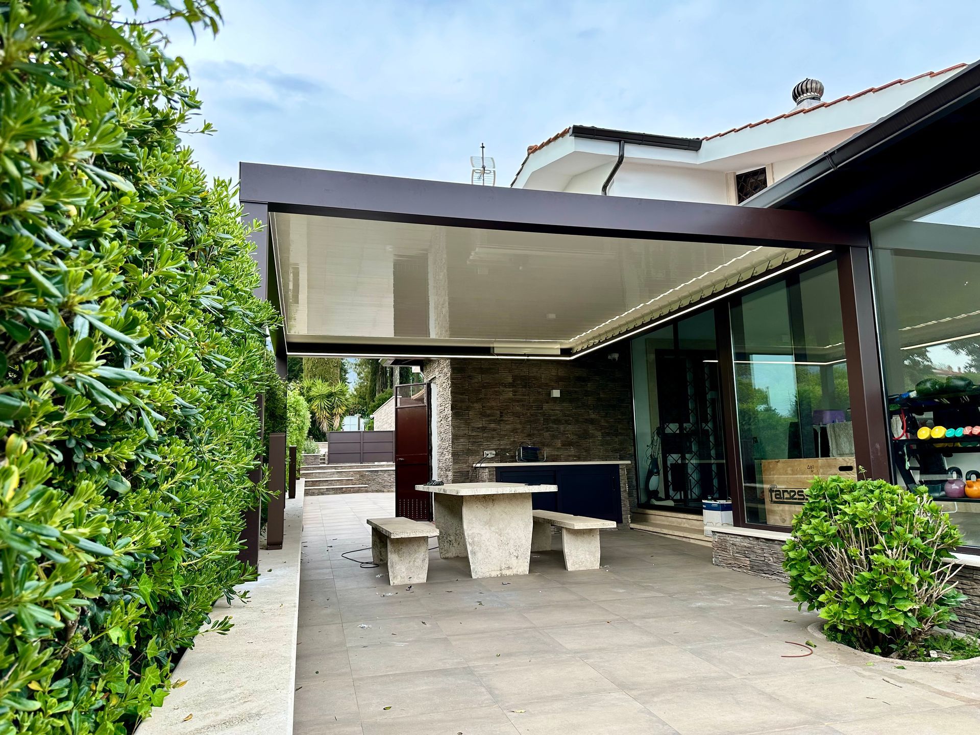 Patio moderno con tavolo in pietra, panchine e zona salotto esterna coperta accanto a un edificio con pareti in vetro.