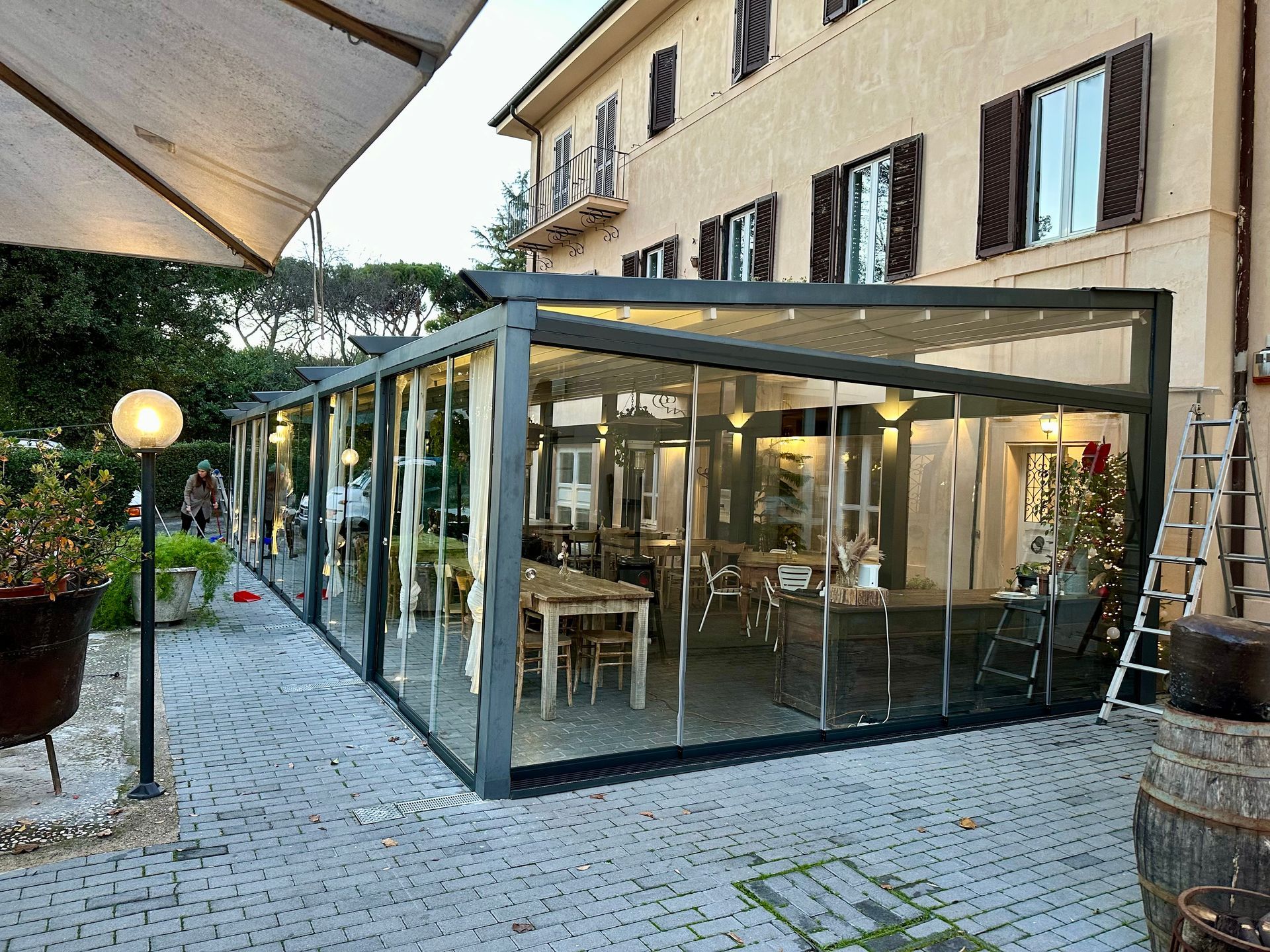 Padiglione-caffetteria con pareti in vetro su una terrazza lastricata di ciottoli accanto a un edificio beige al crepuscolo