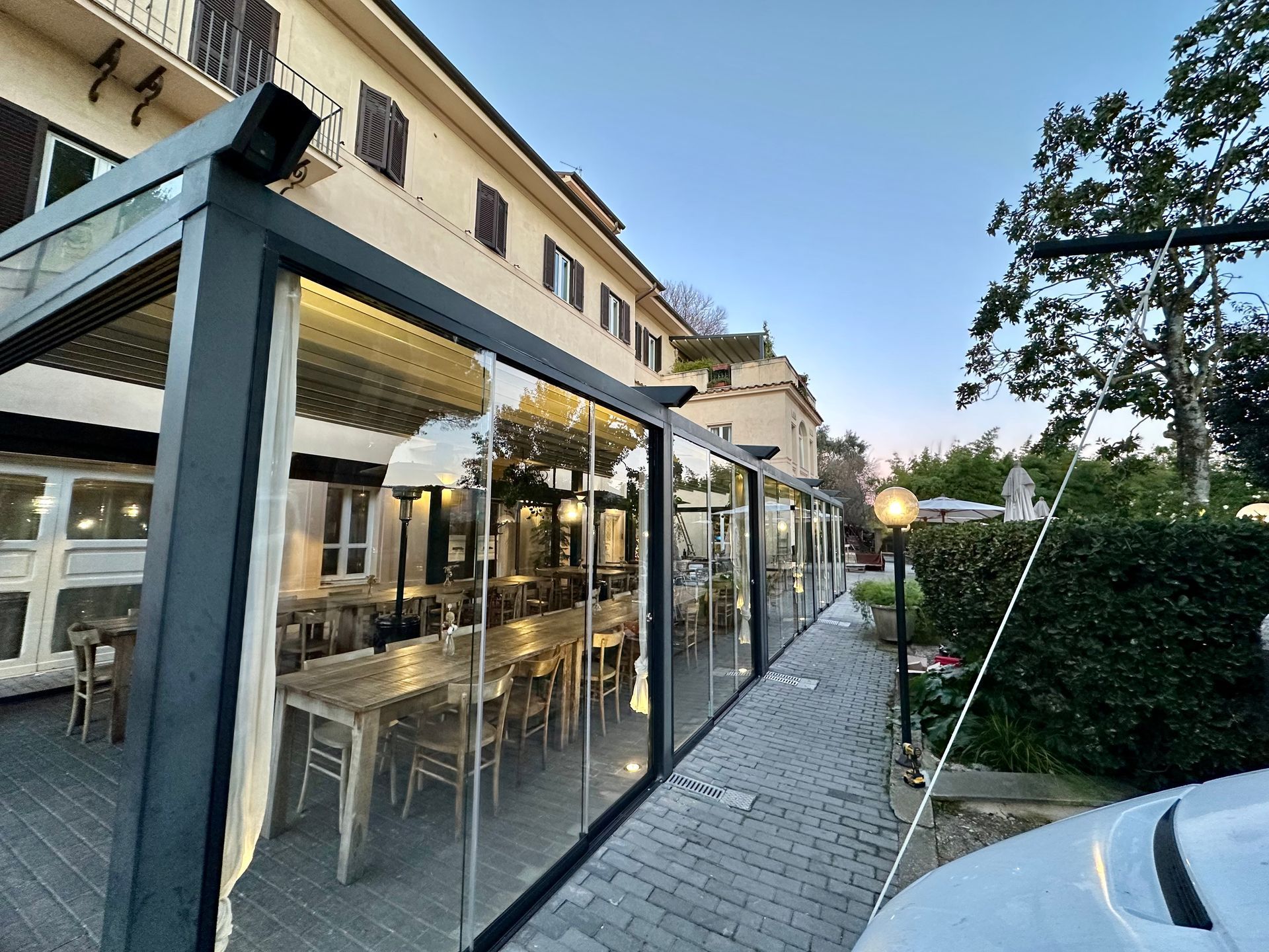 Terrazza di un caffè con pareti in vetro lungo un sentiero lastricato, accanto a una siepe e un'auto parcheggiata al crepuscolo.