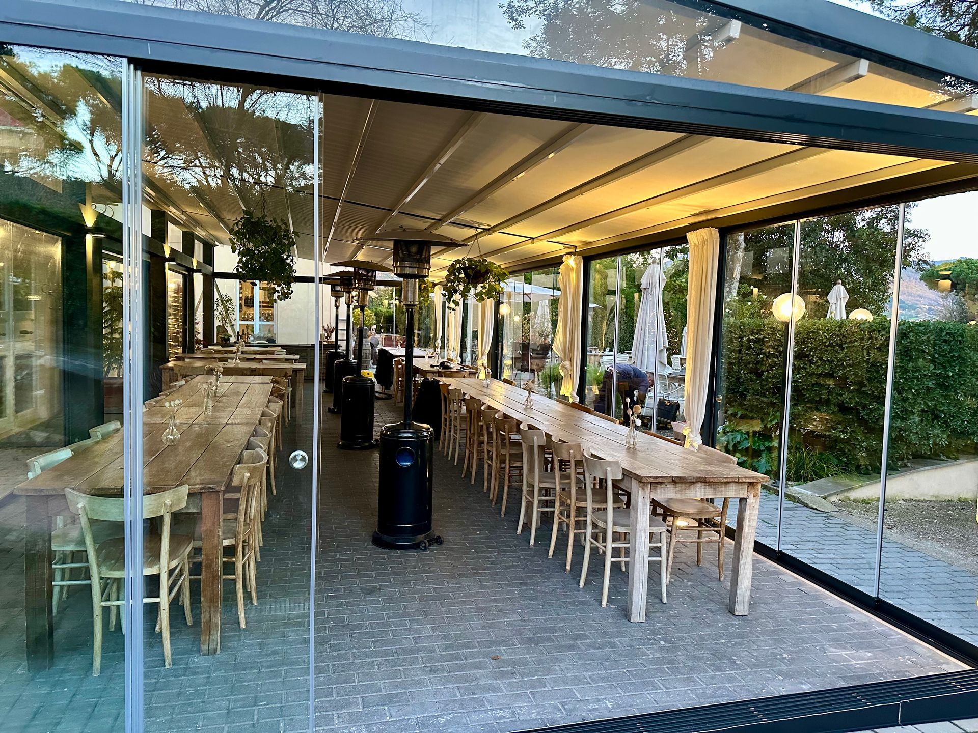Tavolini all'aperto sotto un patio coperto, con lunghi tavoli e sedie in legno lungo una parete di vetro.