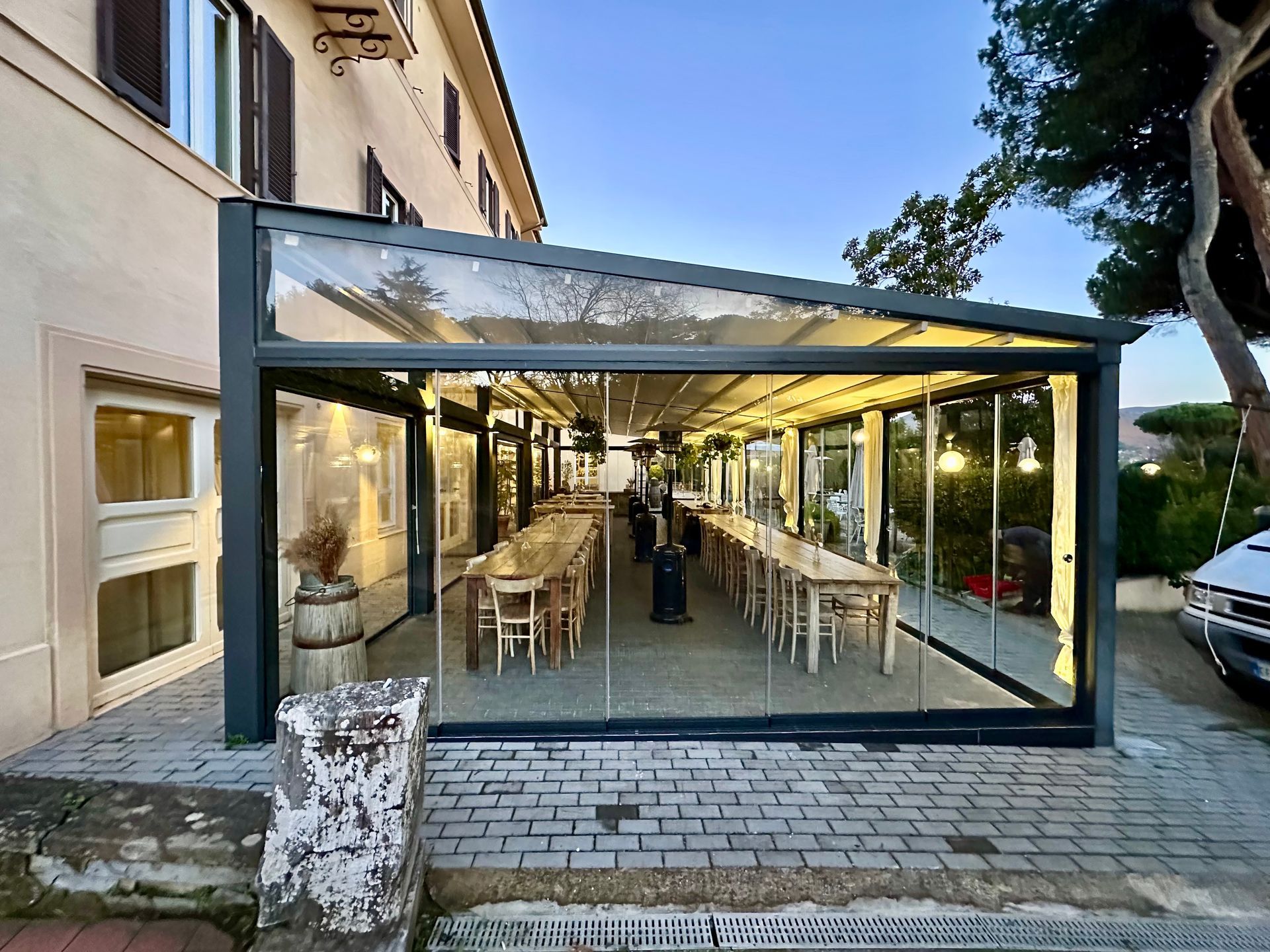 Terrazza esterna di un caffè, racchiusa da vetrate e adiacente a un edificio, con file di tavoli e sedie sotto una tettoia.