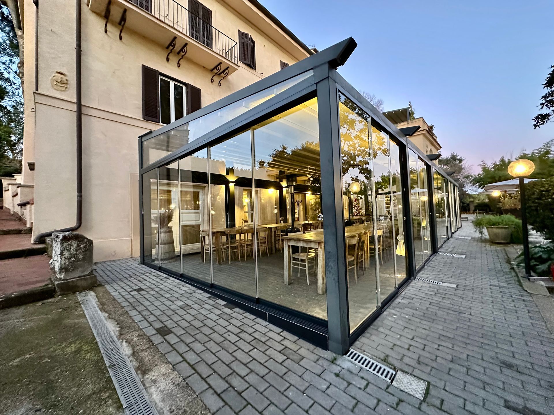 Zona pranzo esterna con pareti in vetro, adiacente a un edificio e illuminata da una luce calda al crepuscolo.