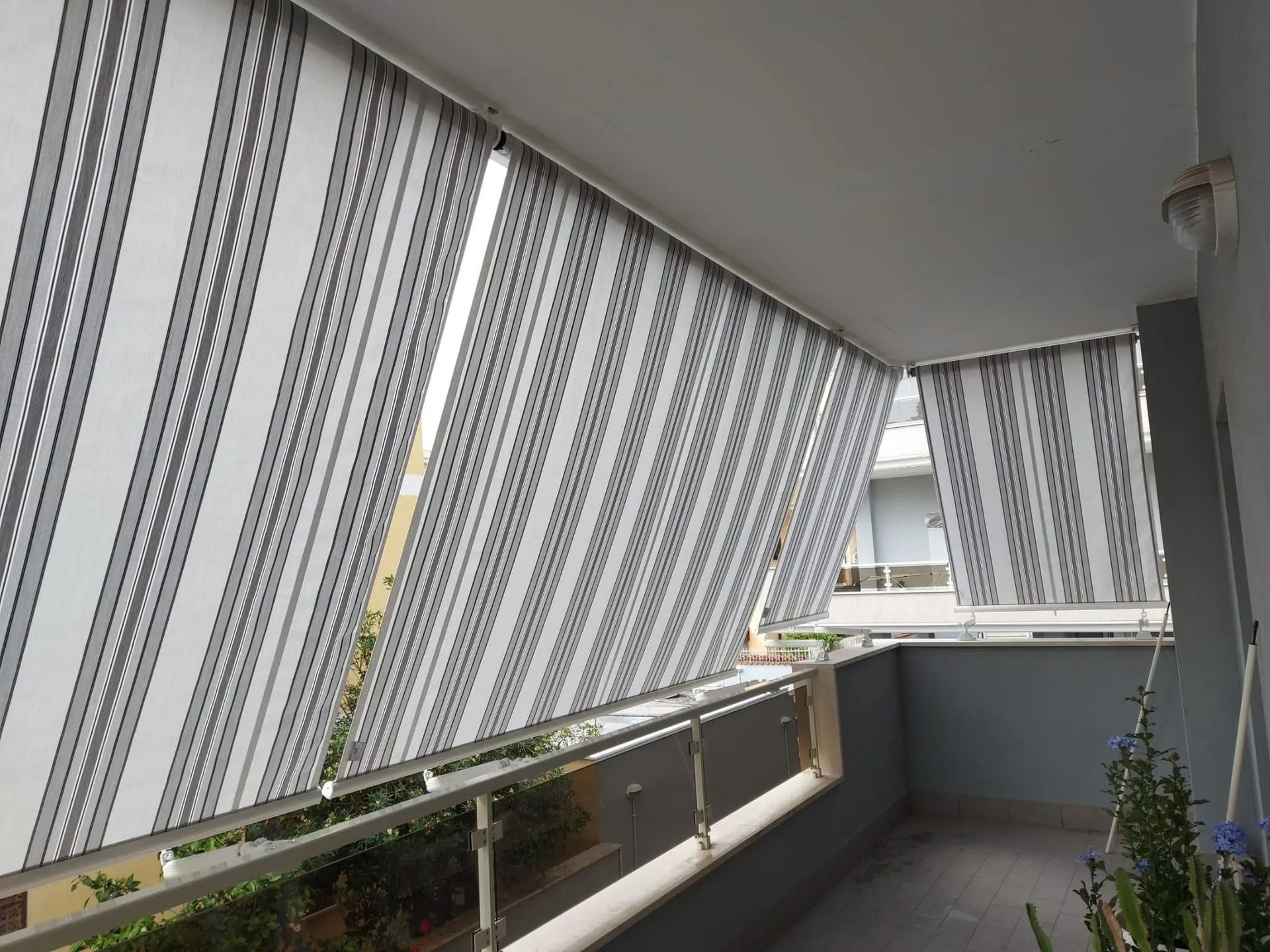 Balcone con tende a righe grigie e bianche, ringhiera in vetro e piante in vaso all'esterno.